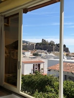 Vista a través de una ventana que muestra un balcón, un muro de piedra y una iglesia distante.