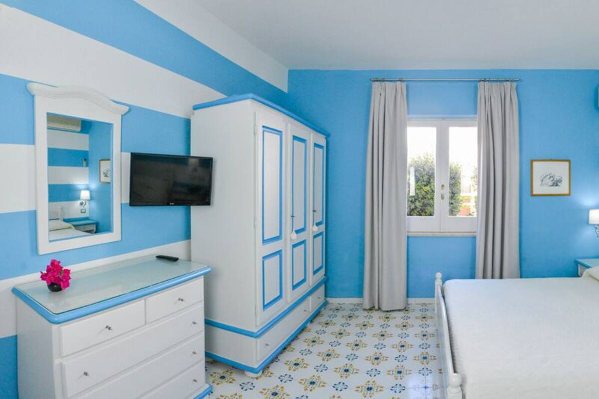 Habitación de hotel con cama, mesita de noche con lámpara, televisión, armario y ventana con cortinas.