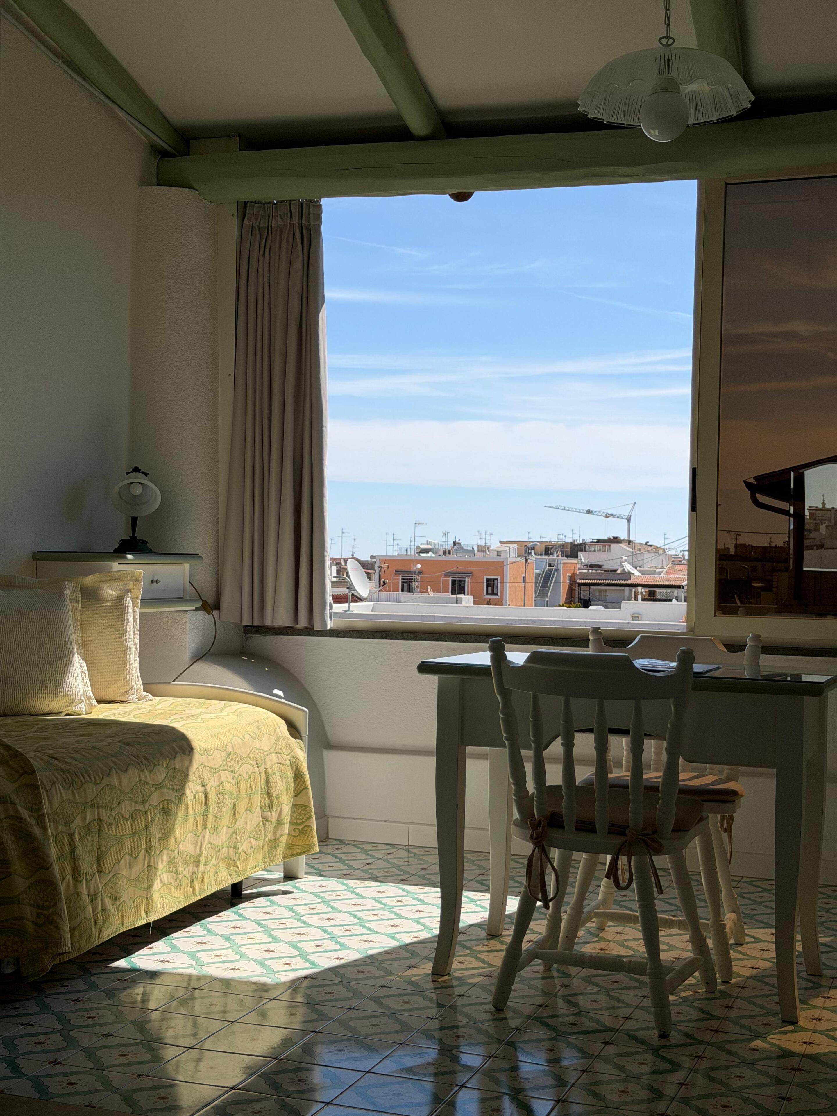 Una habitación con cama, escritorio y silla, una ventana con vistas a edificios y un ventilador de techo.