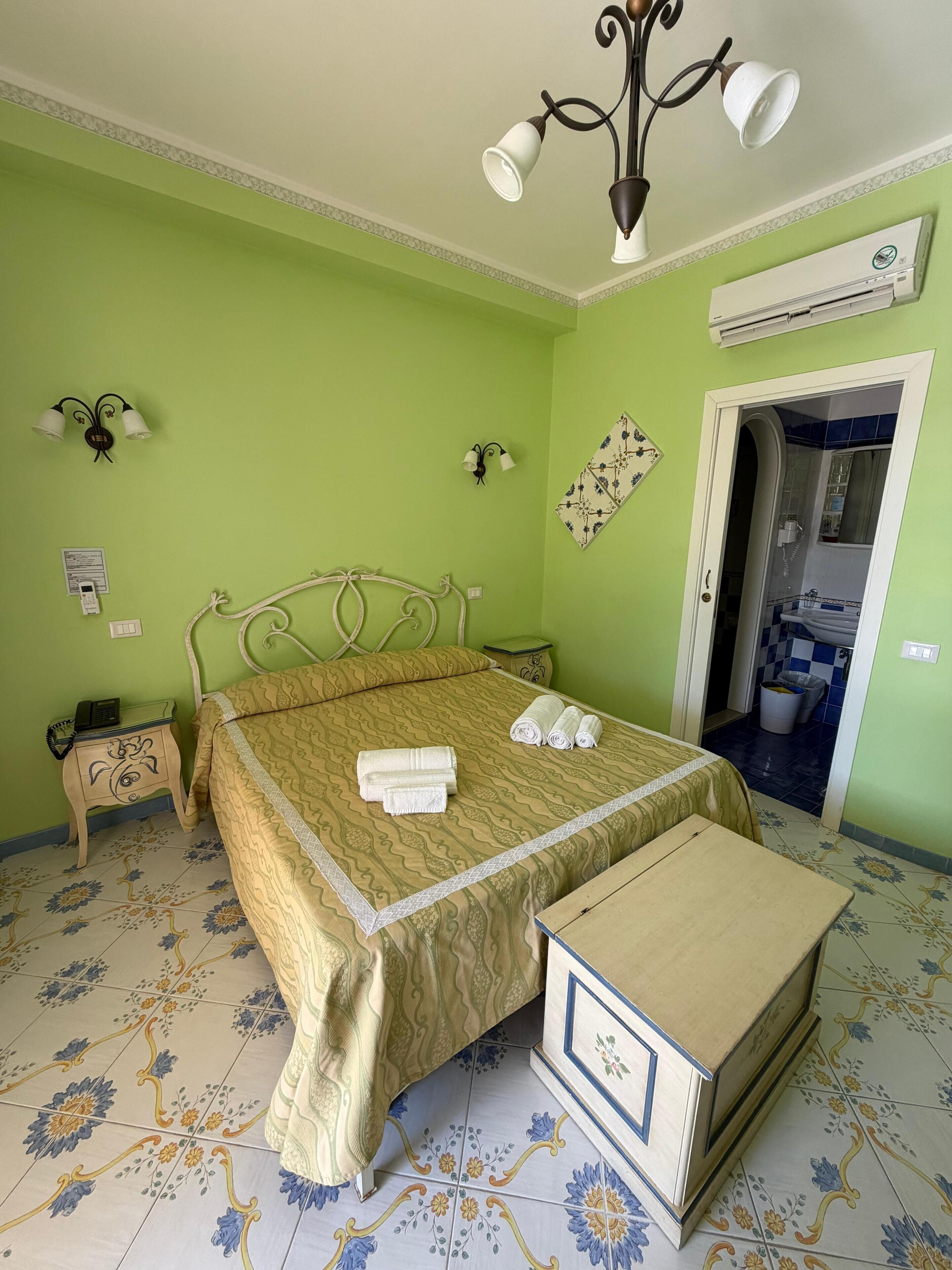 A room with a bed, bedside table, lamp, wall-mounted air conditioner, and a bathroom door.