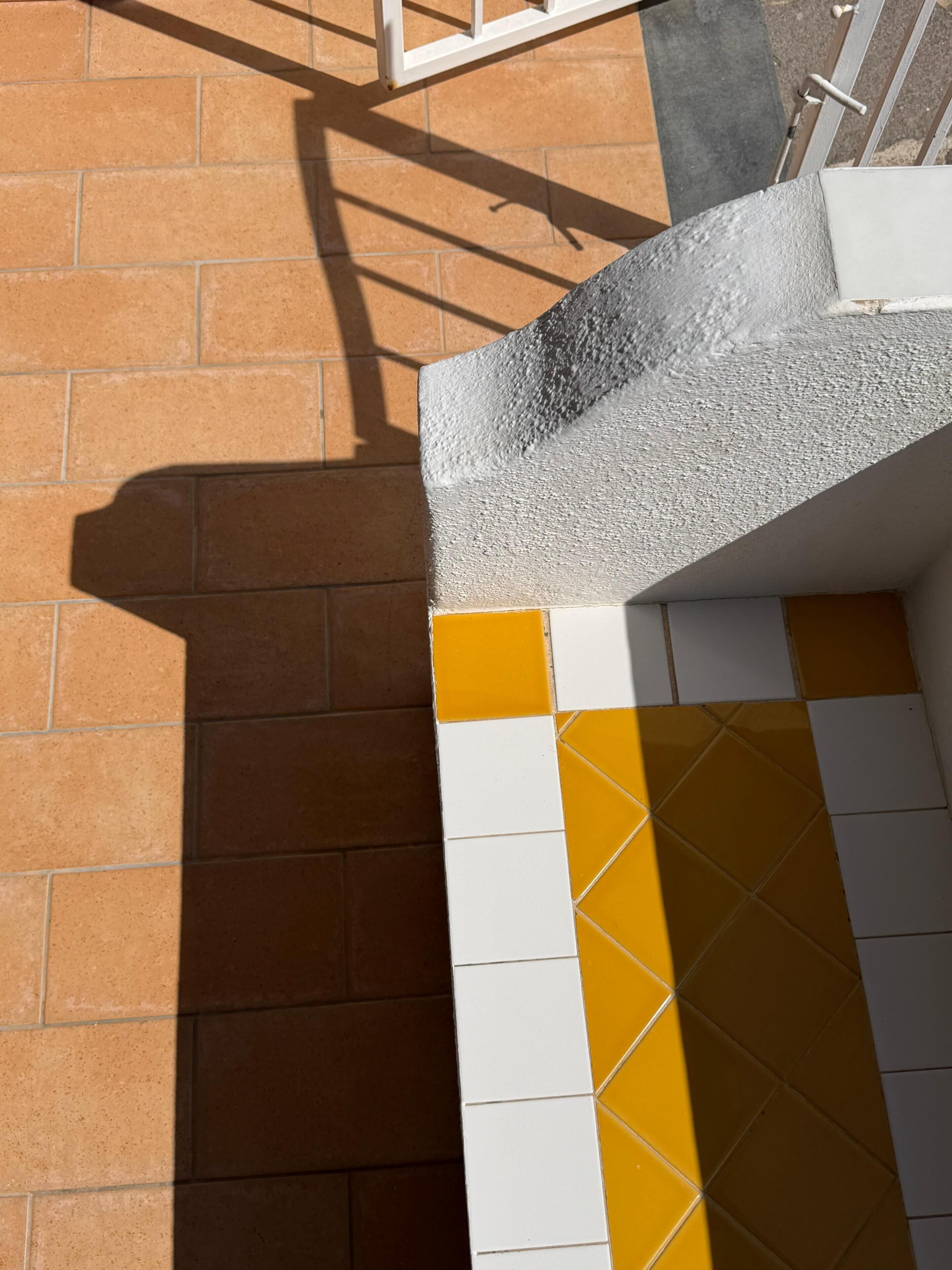 A corner of a building with a mix of brown and white tiles, and a shadow cast on the ground.