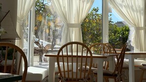 Un espace extérieur aménagé avec des meubles de jardin en bois, composé de chaises et de tables, de grandes fenêtres ornées de rideaux blancs et offrant une vue sur la verdure environnante.