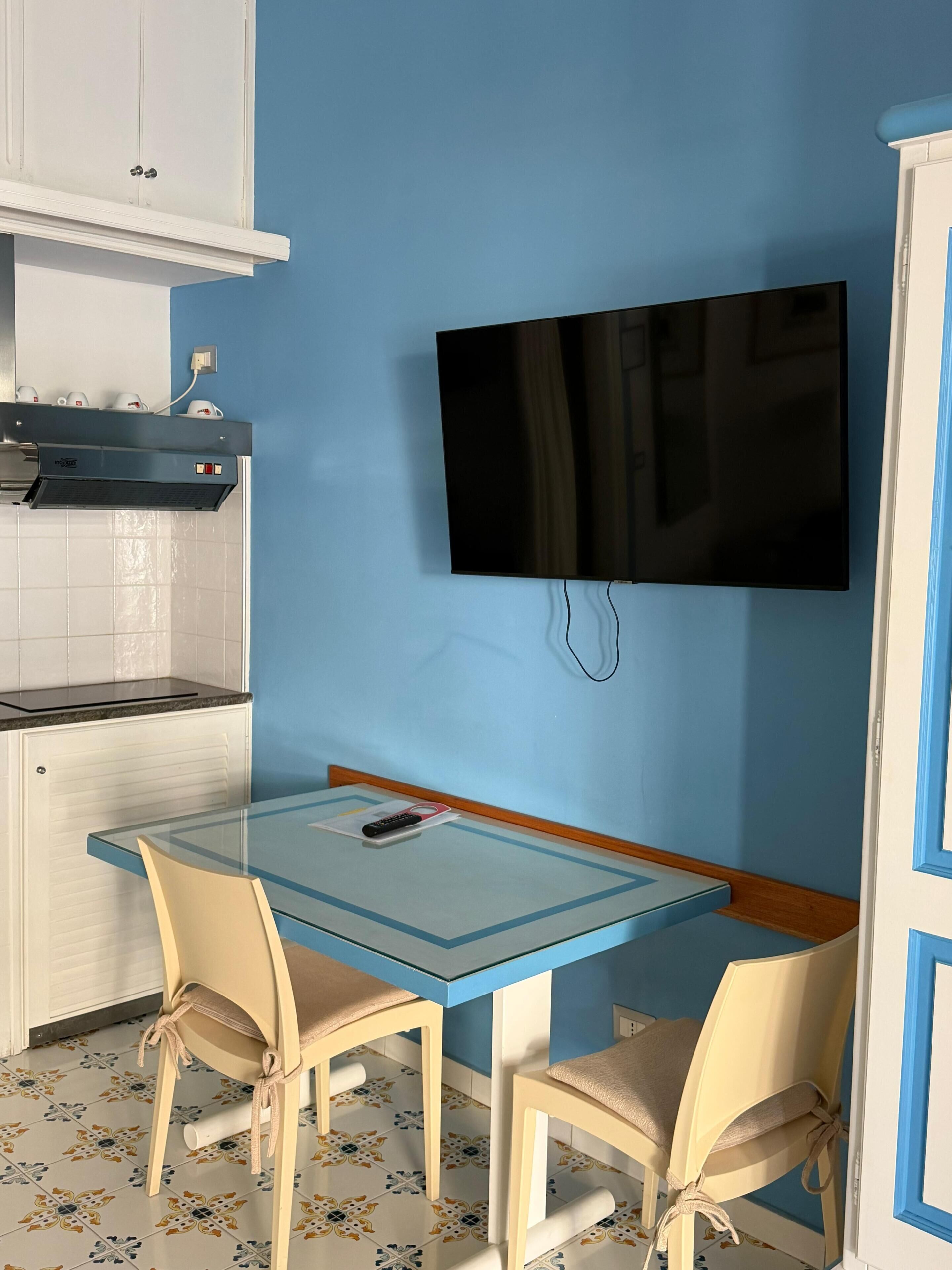 A small kitchen with a TV mounted on the wall, a table with chairs, and a cabinet above the counter.