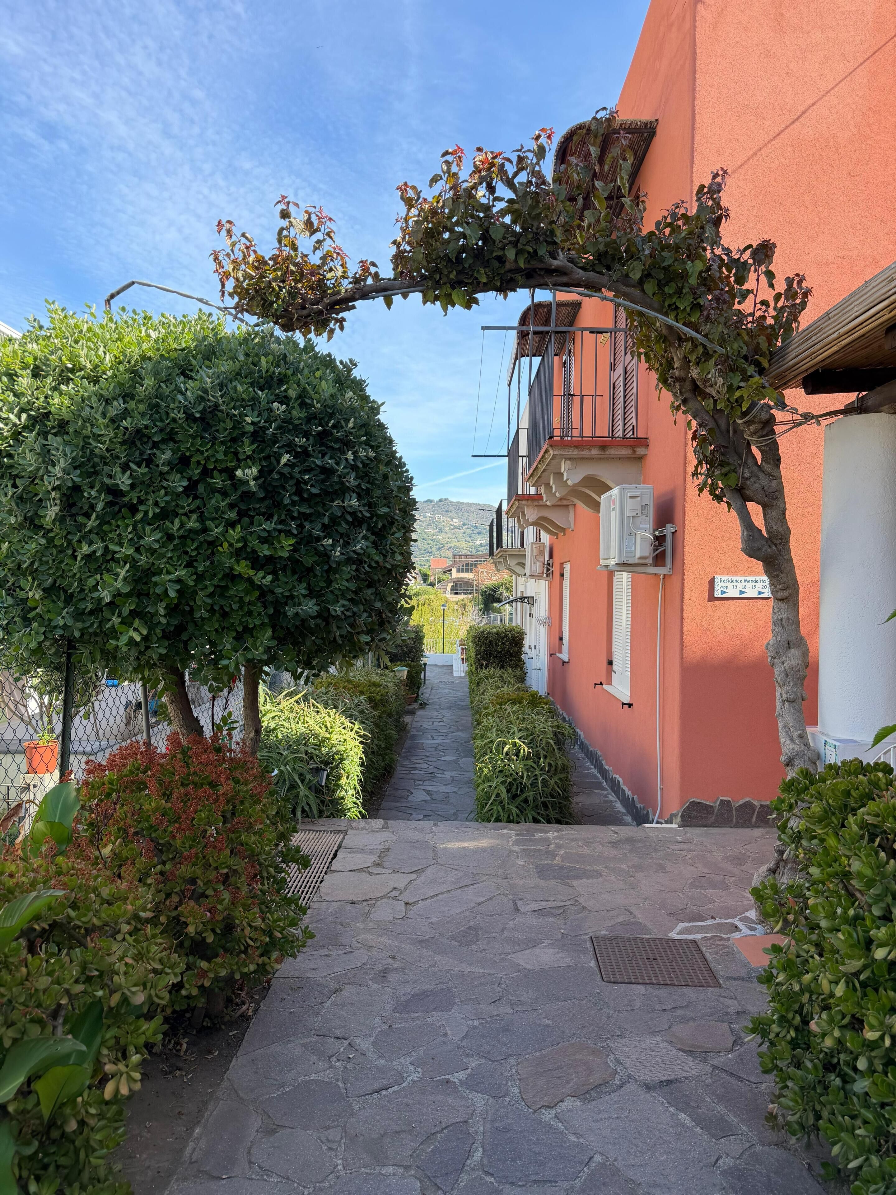 Un étroit sentier de pierres mène à un bâtiment à la façade rouge, entouré de verdure et sous un ciel dégagé.
