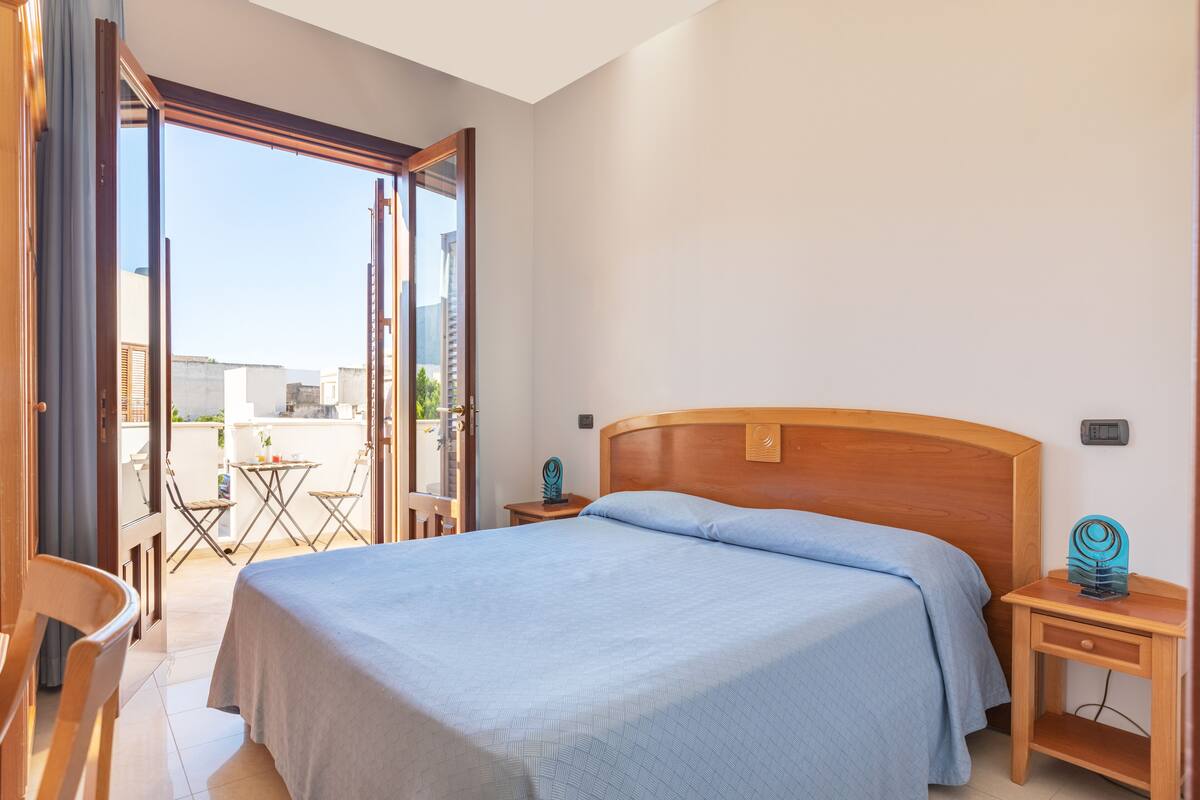 Een hotelkamer met een bed, een houten nachtkastje, een kleine tafel met stoelen en een open deur naar een balkon.