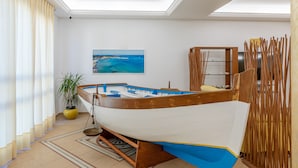 A room with a large window, a potted plant, a wooden boat, and a painting of a beach scene. - Hotel Sabbia D'Oro (San Vito Lo Capo)