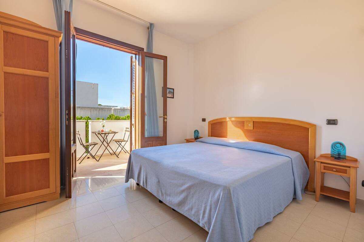 Een slaapkamer met een bed, een houten nachtkastje, een kleintje tafeltje met stoelen, en uitzicht buiten.