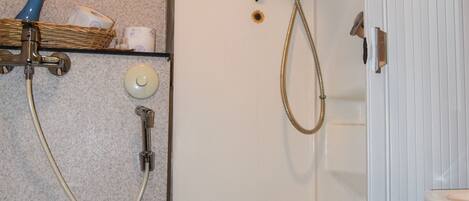 A bathroom with a toilet, a shower with a hose, a framed botanical print on the wall, and a small shelf with toiletries.