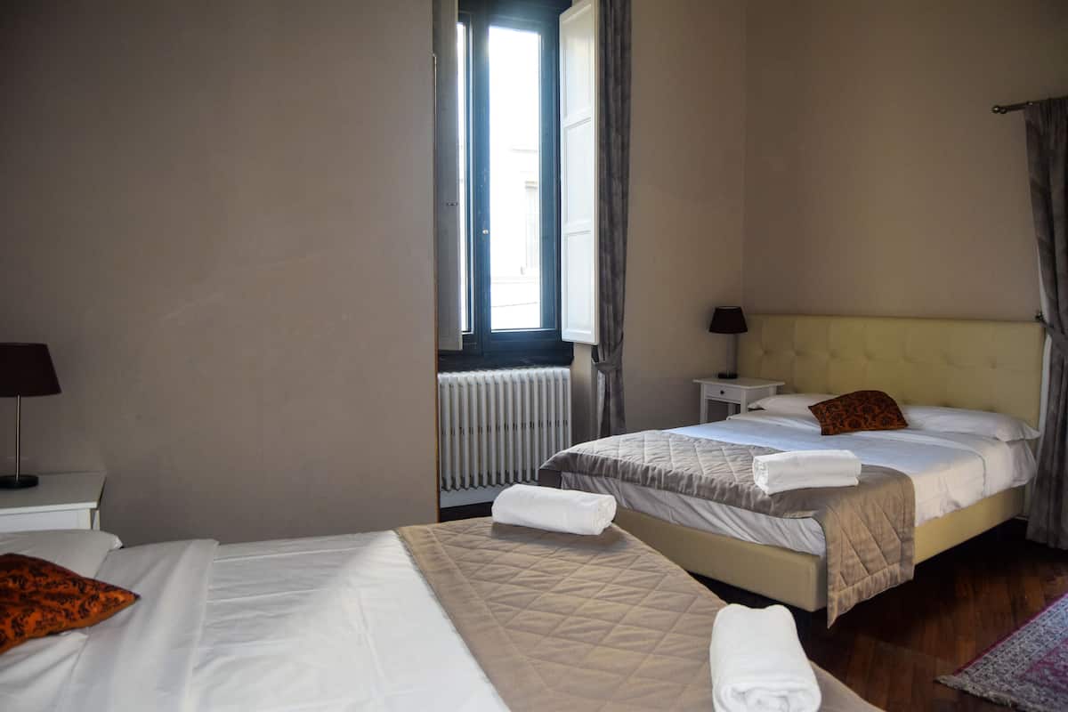 A hotel room with two beds, a window with curtains, a radiator, and a bedside table with a lamp.