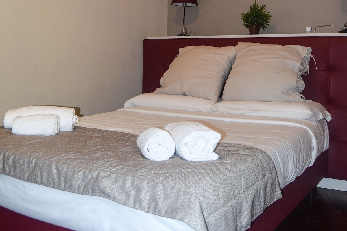 A neatly made bed with white linens and two rolled towels, a red headboard, a framed picture of a building on the wall, and two bedside lamps.