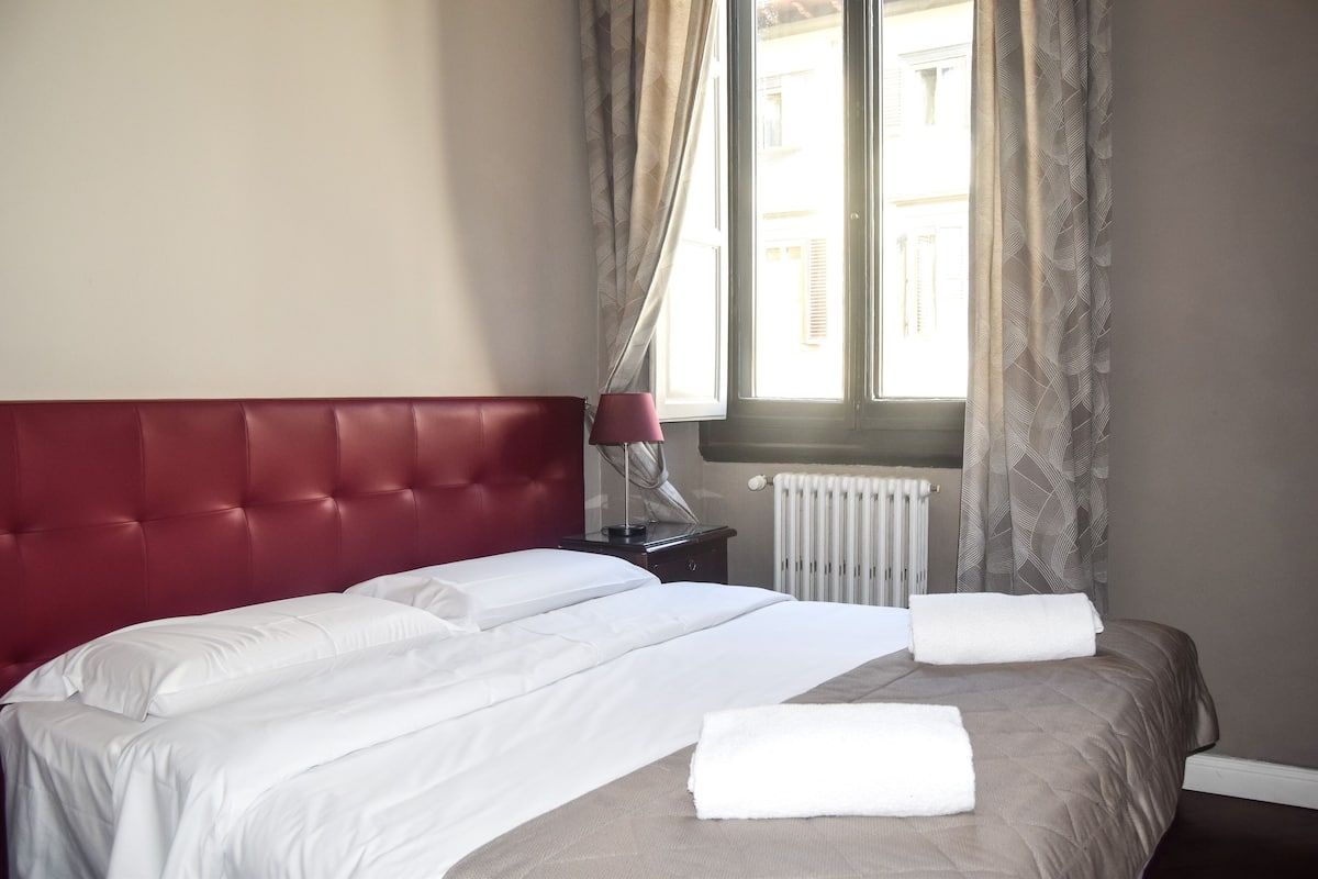 A neatly made bed with white linens, a red headboard, and a window with curtains.