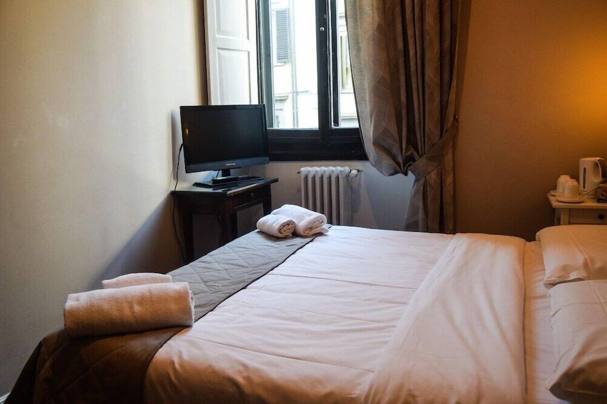 A hotel room with a bed, a television on a desk, a window with curtains, and a radiator.