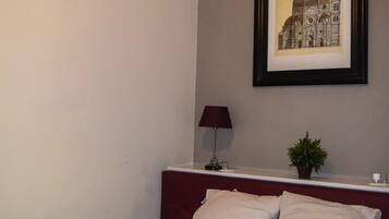 A neatly made bed with white linens and pillows, a framed picture of a building on the wall, a small table with a lamp, and a potted plant.