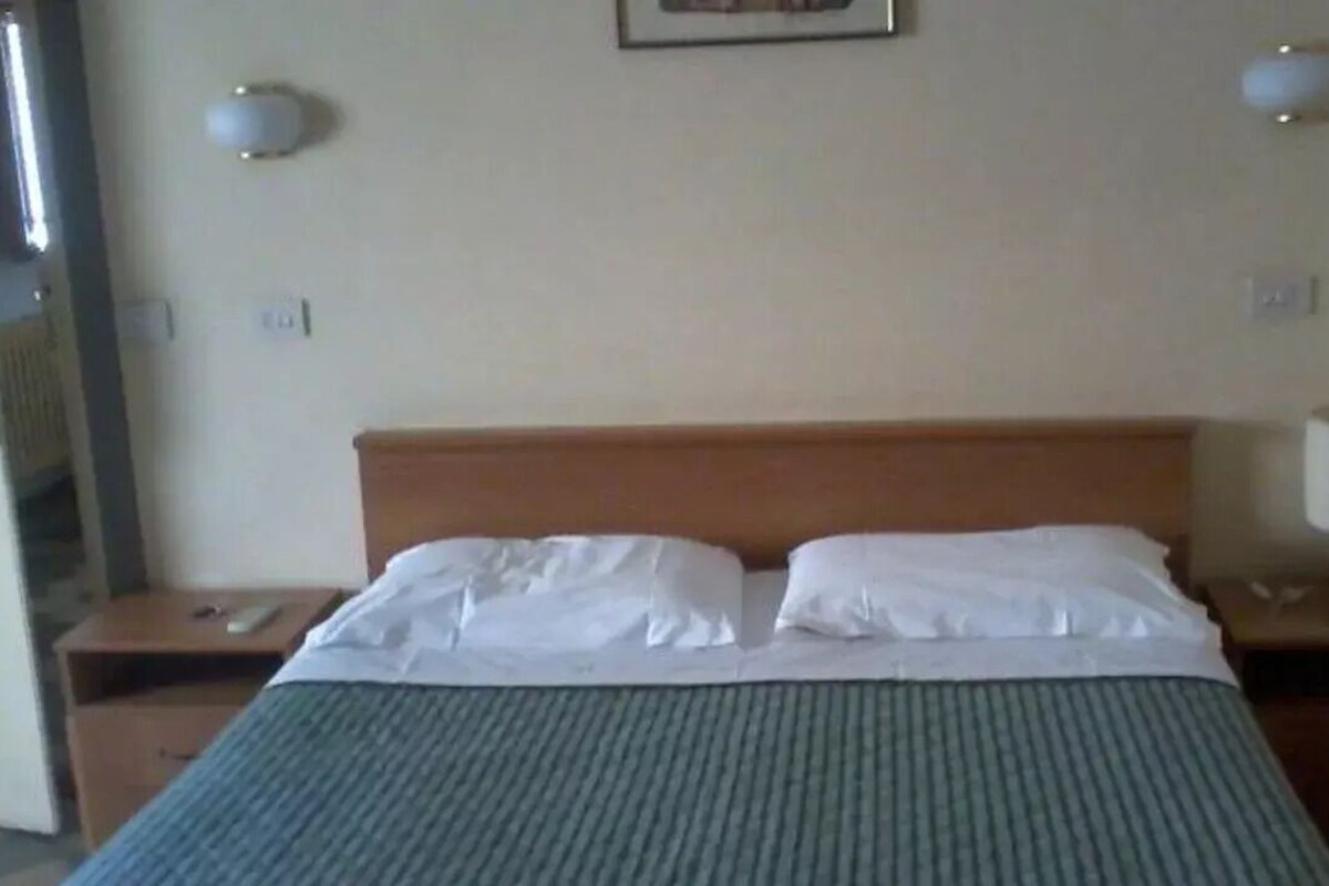 A hotel room with a bed, two bedside tables, a nightstand, a lamp, and a wall-mounted light fixture.