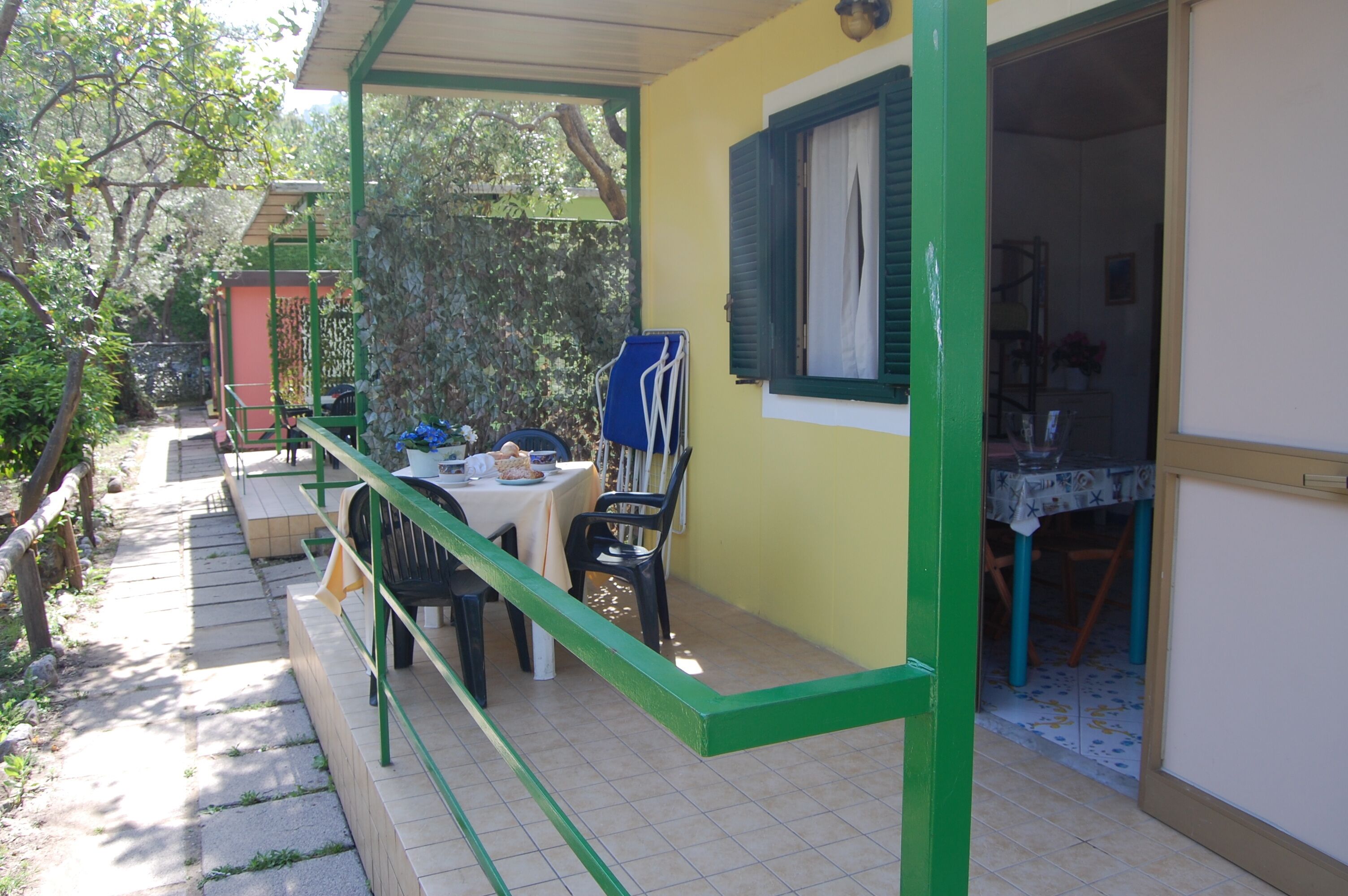 Un espacio de comedor al aire libre cubierto con una mesa y sillas, baranda verde y pared amarilla con una ventana.
