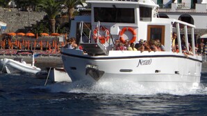 Un barco llamado 'Nerano' con pasajeros, un flotador salvavidas y una costa rocosa con sombrillas al fondo.