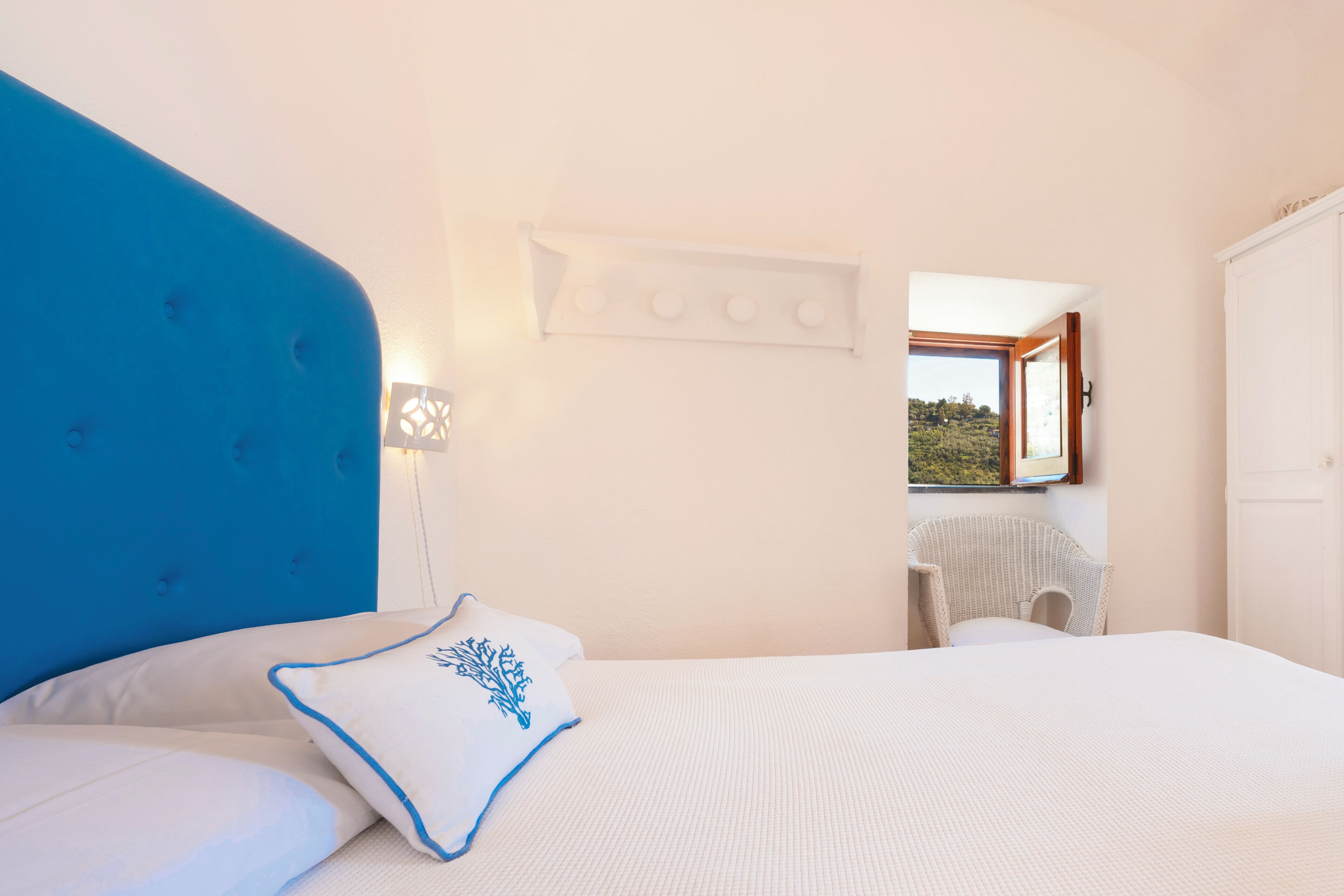 A bedroom with a blue headboard, a white bedspread, and a white pillow with a blue design. There is a white wall-mounted light fixture and a window with a view of greenery outside.