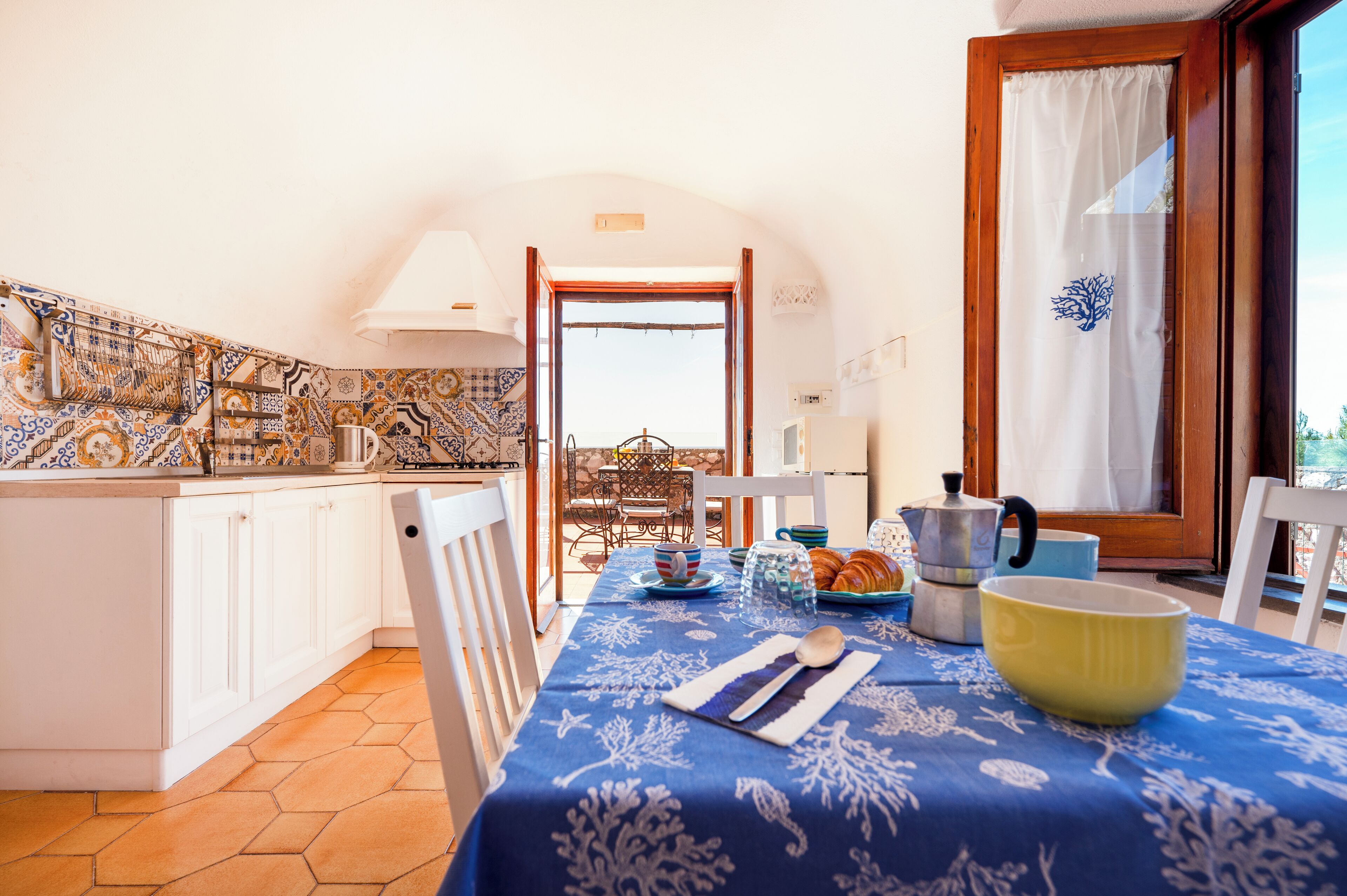 A kitchen with a blue tablecloth, a coffee pot, and a window with a curtain.
