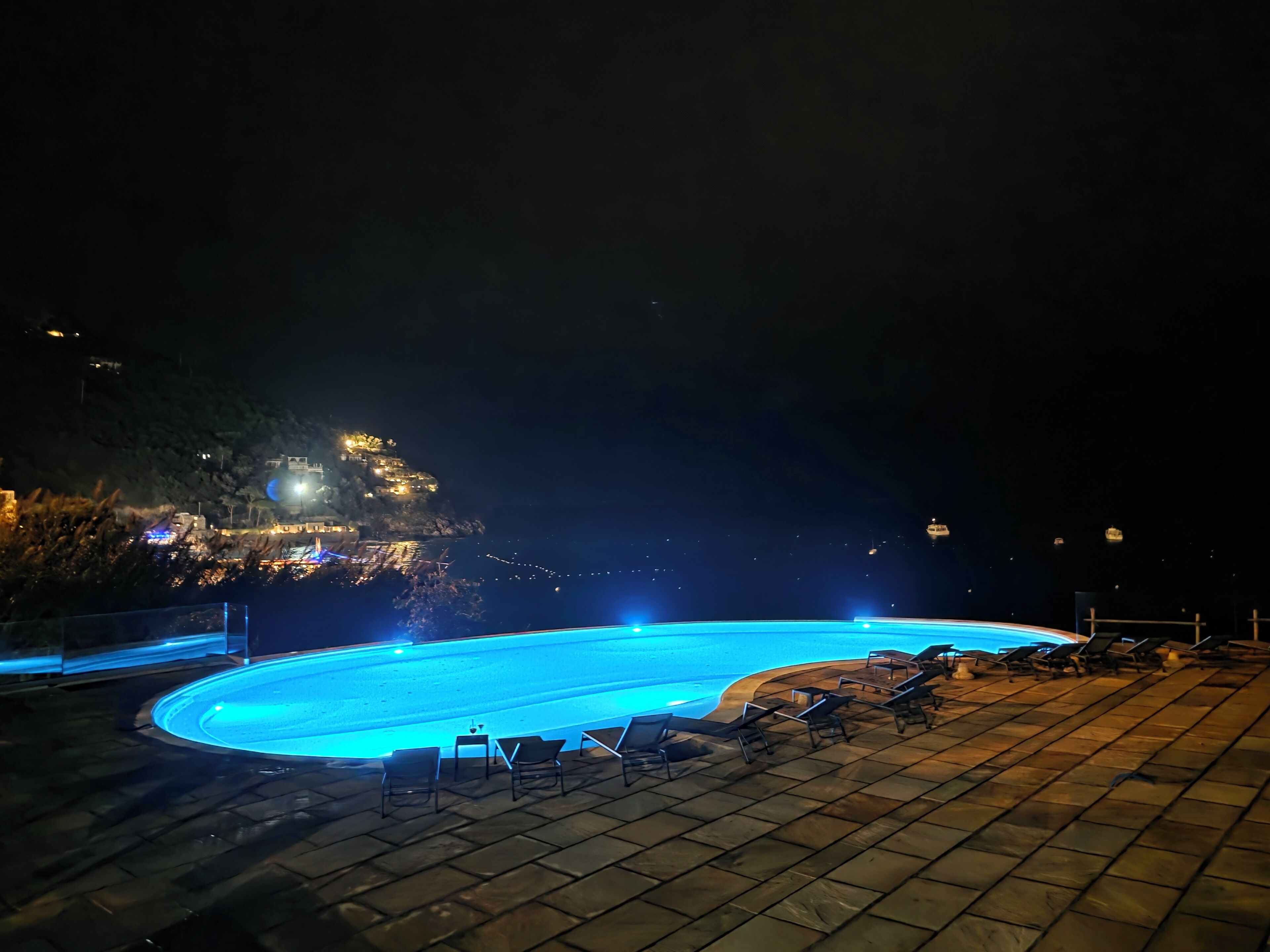 A nighttime swimming pool area with lounge chairs and a view of a hillside with lights.