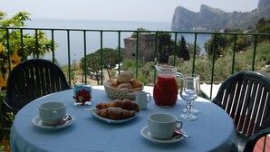 Mesa con croissants, una jarra de jugo rojo y tazas, con vista panorámica a un cuerpo de agua y acantilados rocosos.