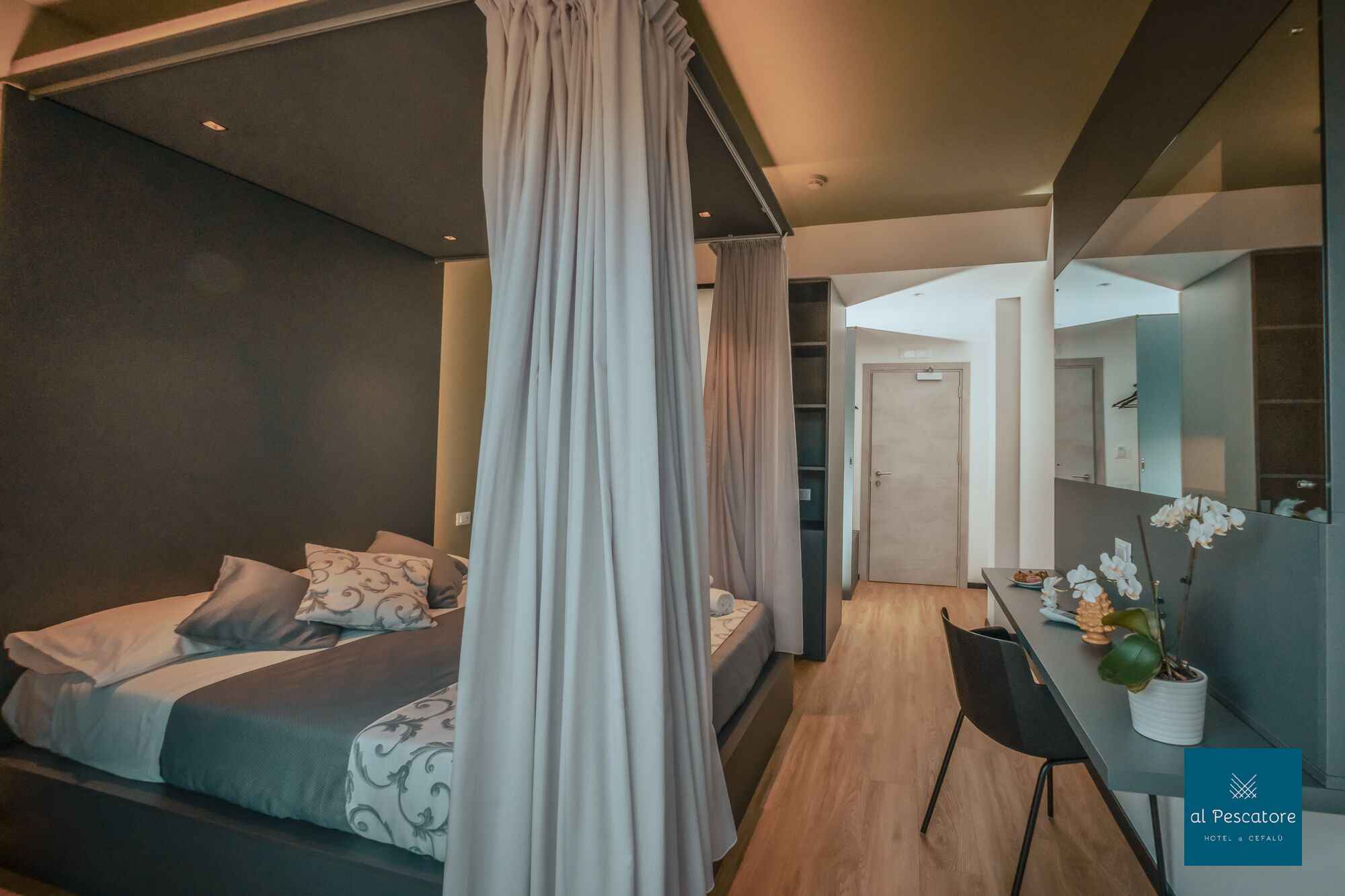 A modern bedroom with a canopy bed, a small desk with a potted plant, and a mirror reflecting the room.