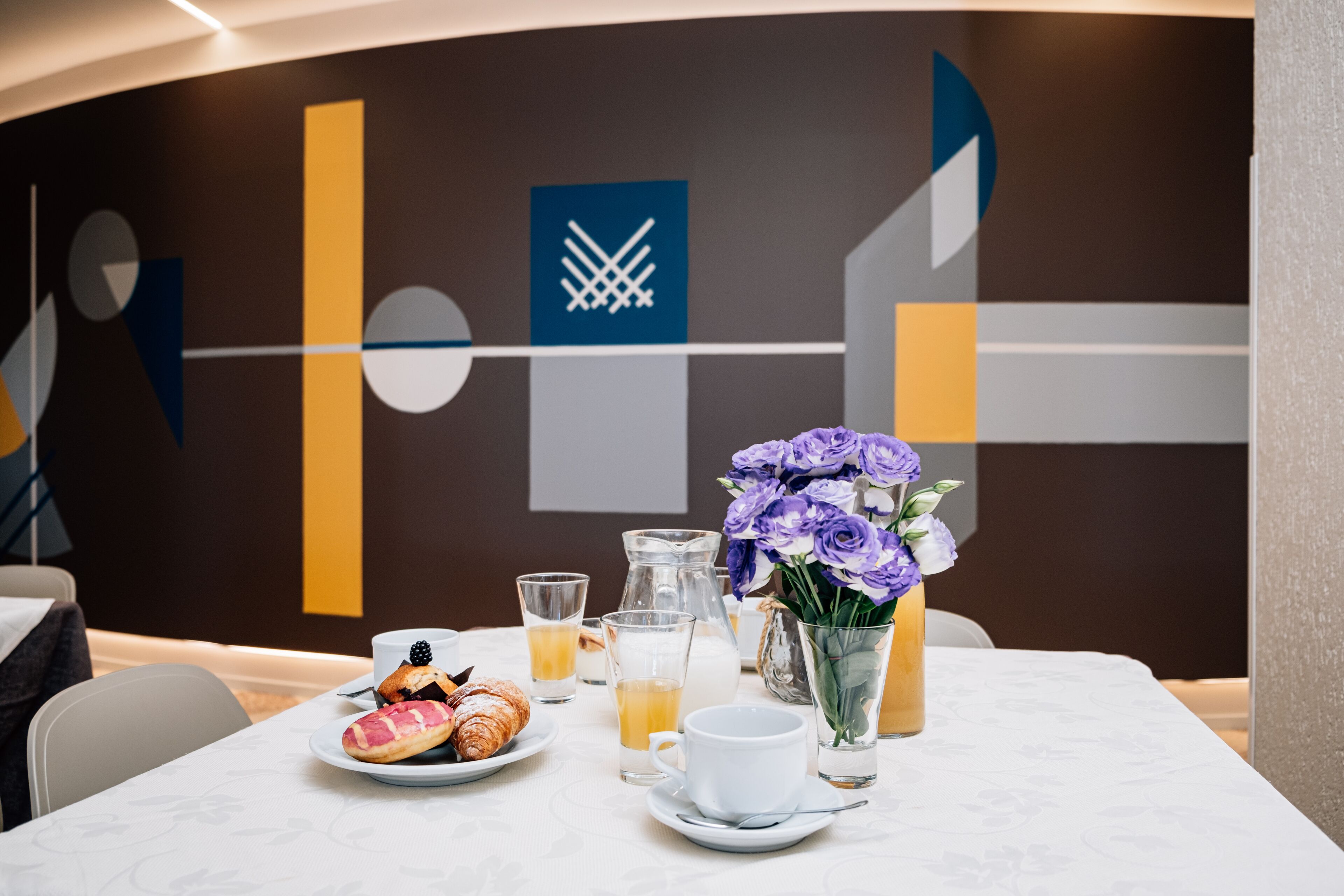 Desayuno con cruasanes, una jarra de jugo de naranja, una taza y un florero con flores moradas sobre una mesa.