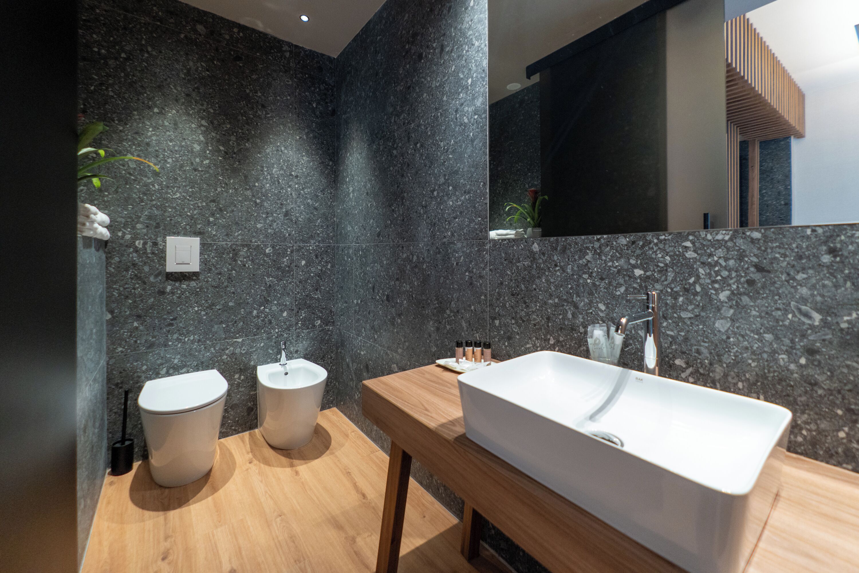 A modern bathroom with dark marble walls, two toilets, a wooden vanity, and a large sink.