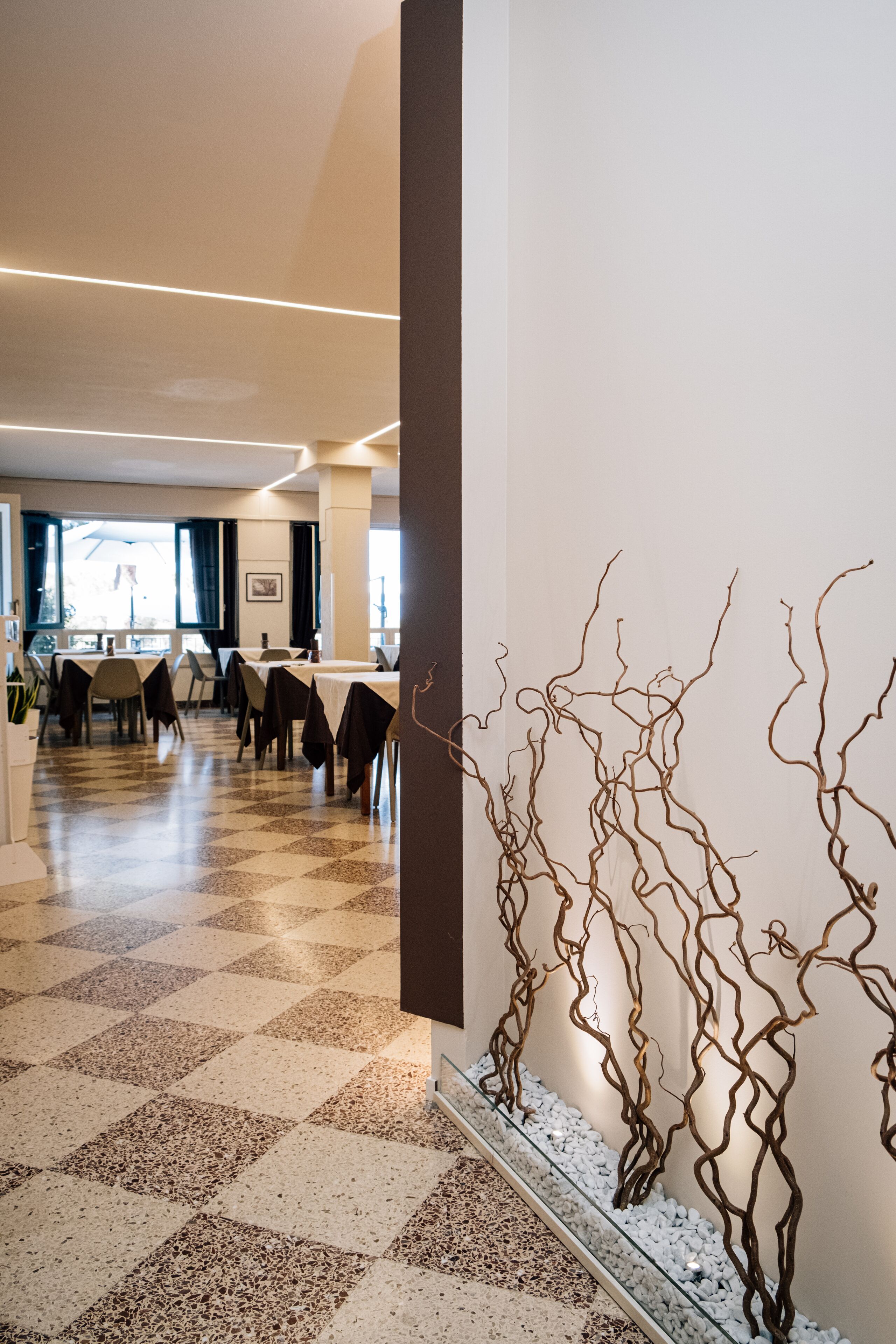 A hotel lobby with a checkered floor, a glass railing with decorative branches, and tables and chairs arranged for dining.