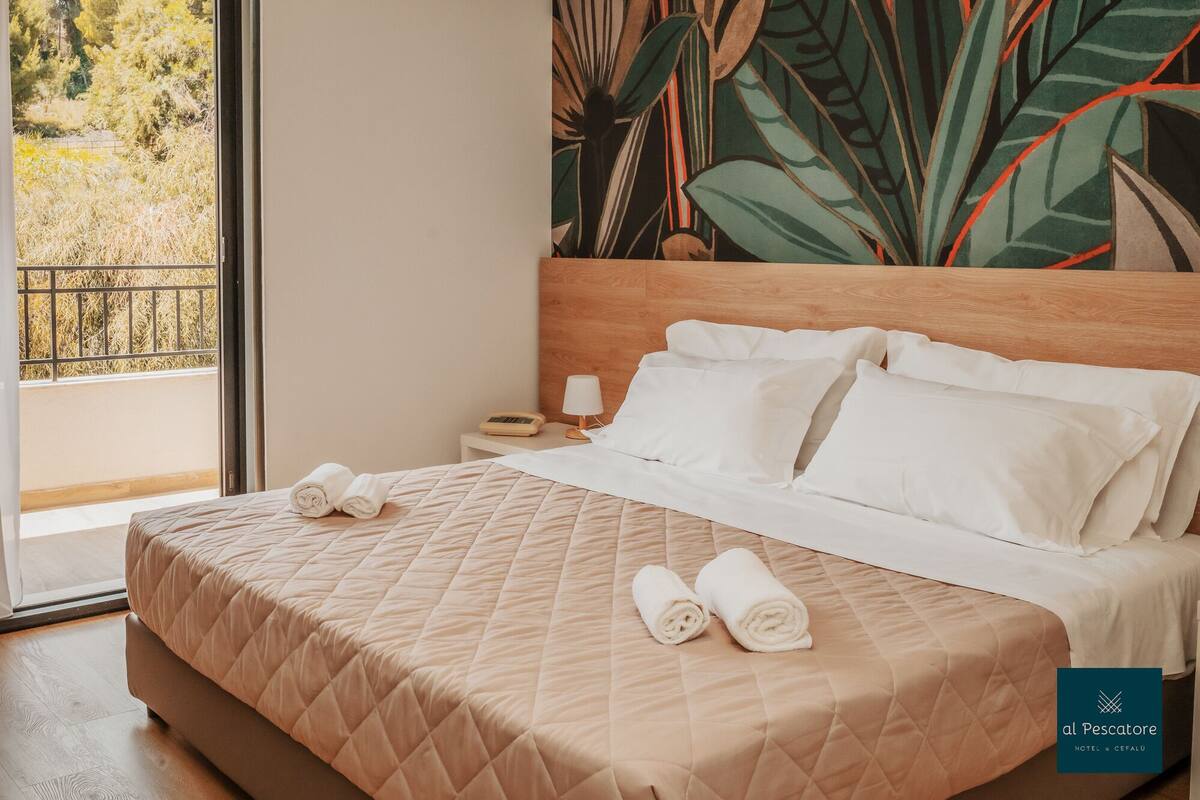 Ein Hotelzimmer mit Bett, Nachttisch, Lampe und Blick ins Freie.