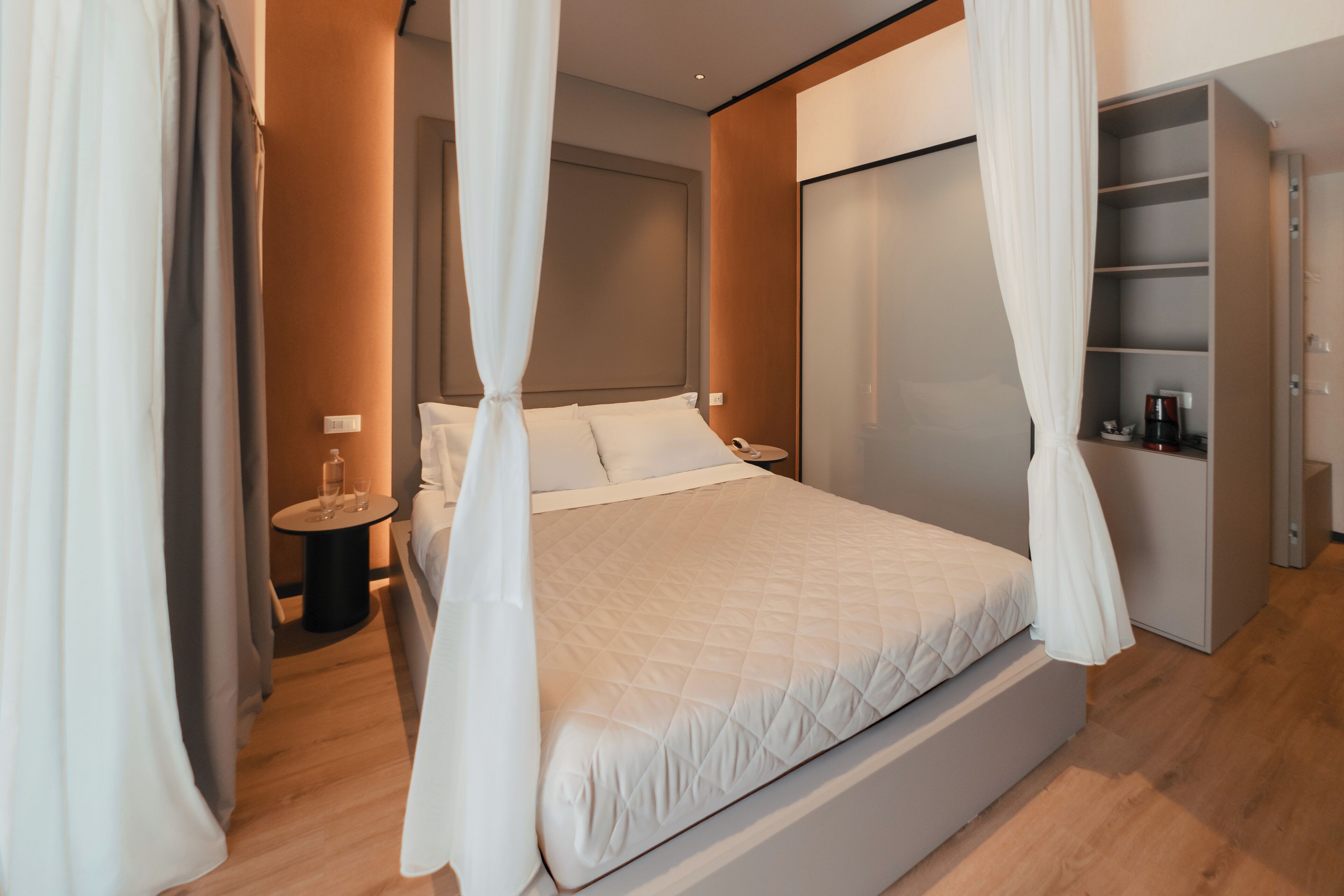 A modern bedroom with a large bed, a bedside table with a lamp, a wardrobe, and a small table with a bottle and glasses.