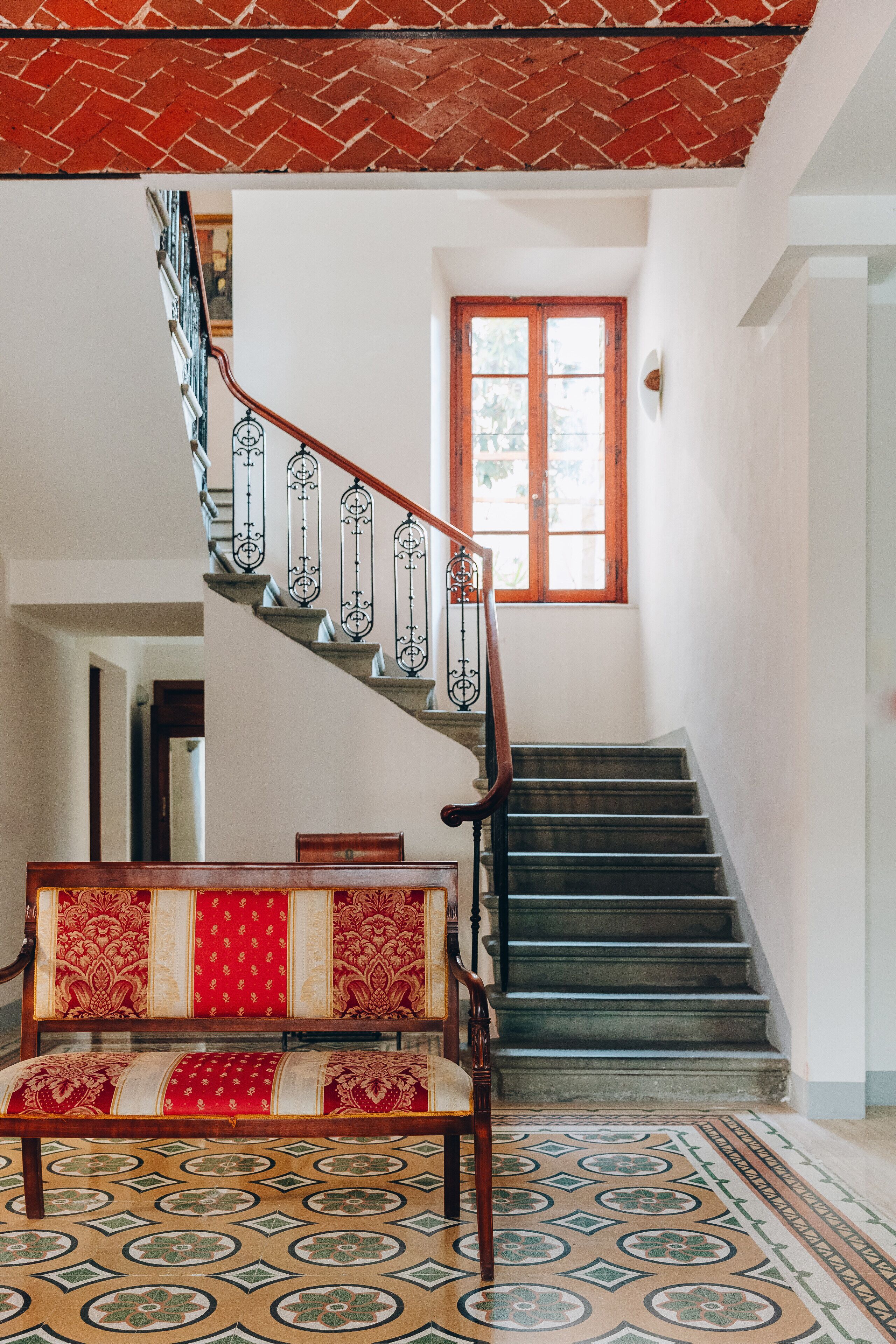 Una escalera con techo de ladrillo rojo y alfombra estampada que conduce a un banco de madera con tapicería roja.