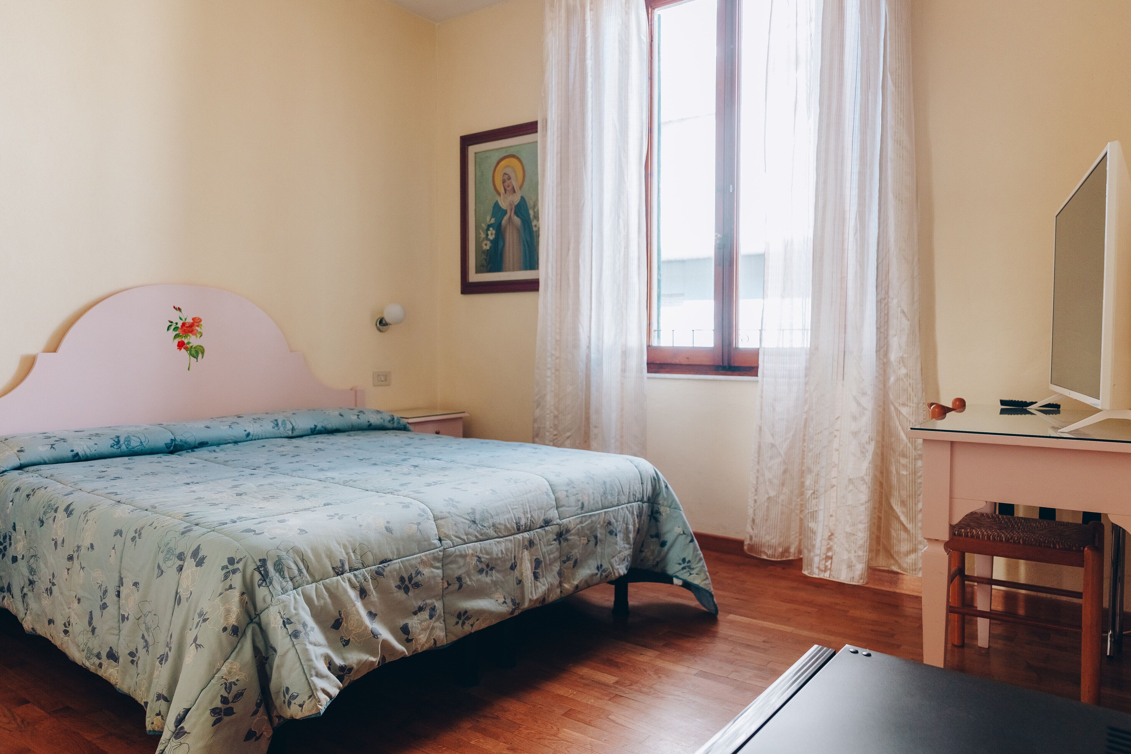 A bedroom with a bed, a desk, a chair, a window with curtains, and a painting on the wall.