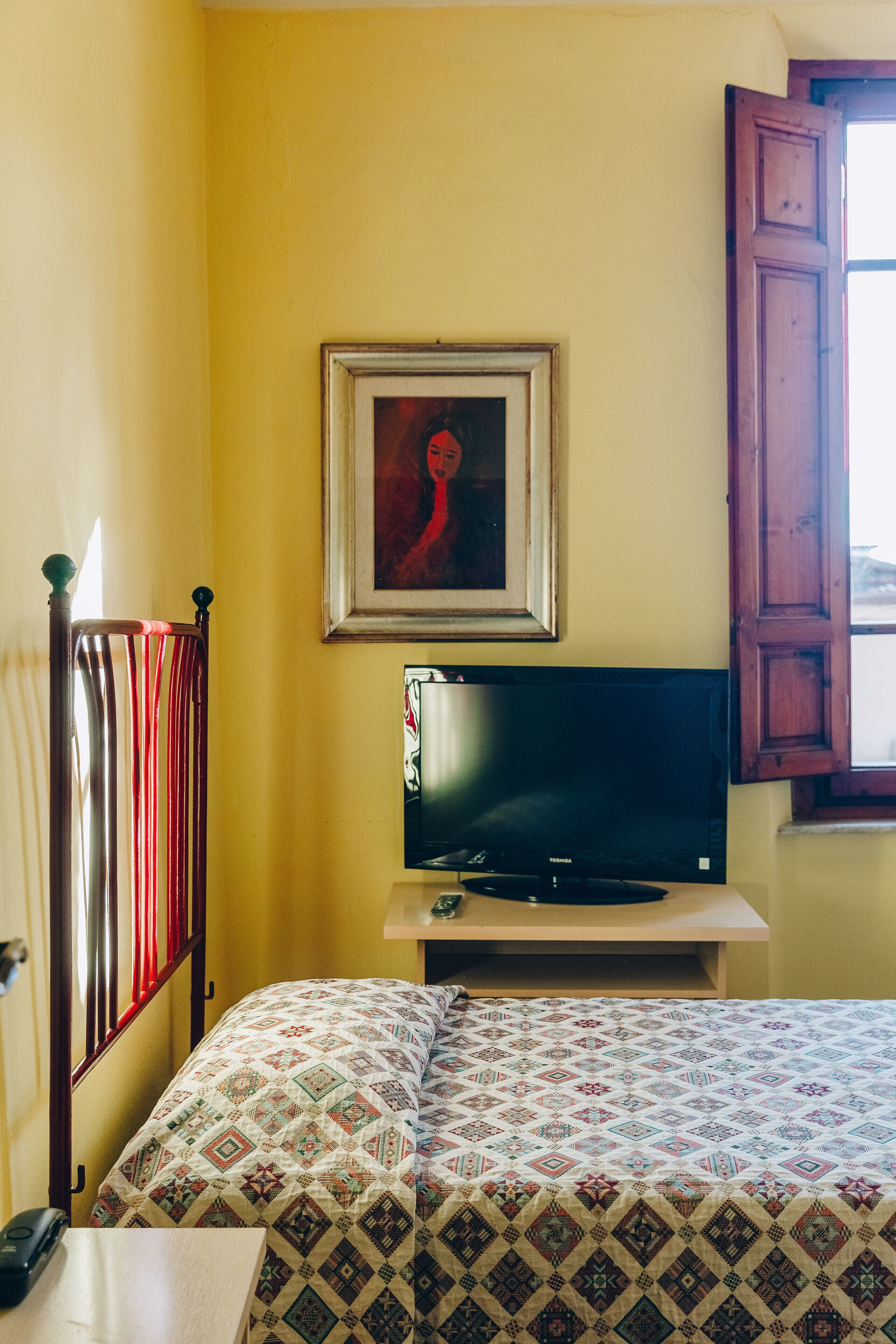 Una habitación con una cama, un televisor sobre un mueble bajo, un cuadro en la pared y una ventana con un persiano de madera.