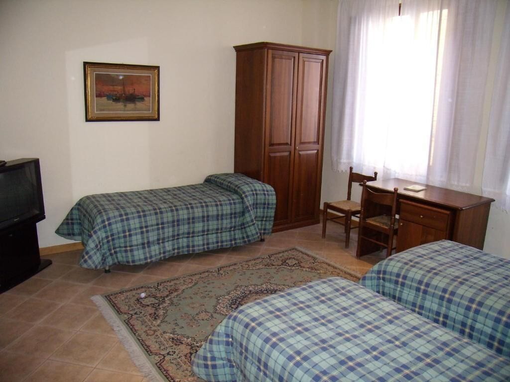 A room with two beds, a wooden wardrobe, a desk with a chair, a television, and a painting on the wall.