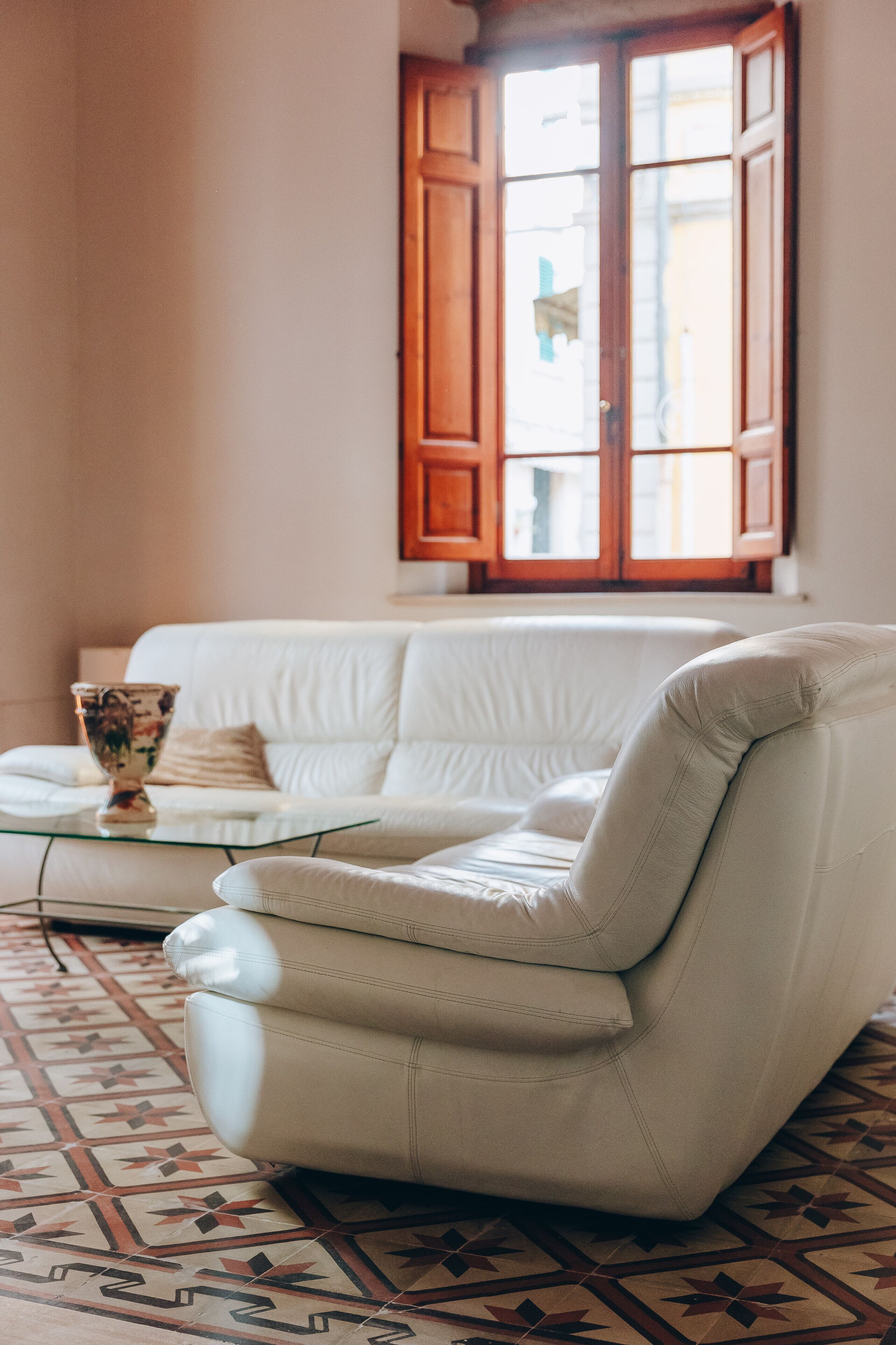 Ein weißes Leder-Sofa und ein Sessel in einem Zimmer mit gemustertem Boden und einem Fenster mit Holzjalousien.