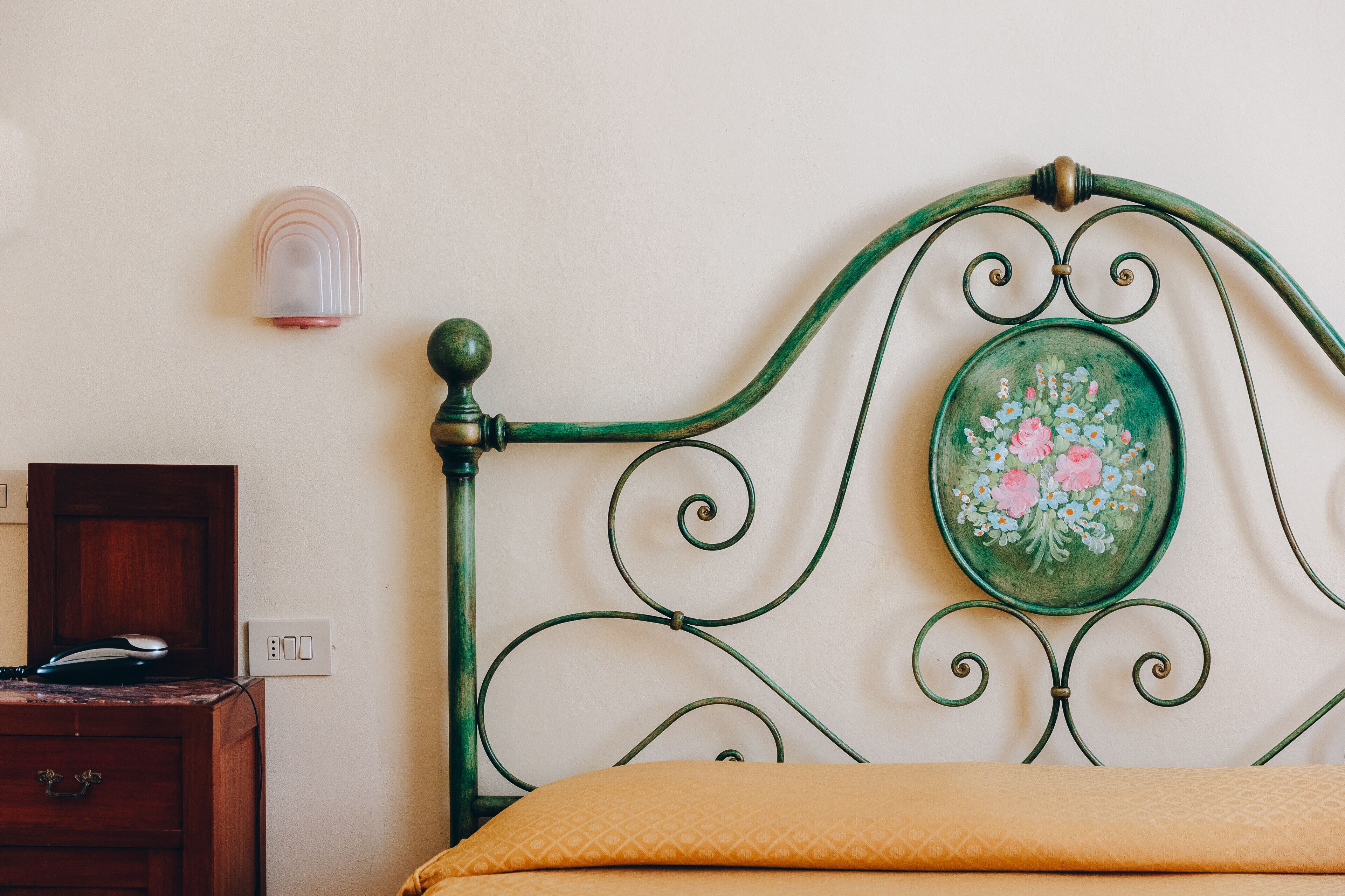 A bed with a green metal headboard, a wooden nightstand, and a wall-mounted light fixture.