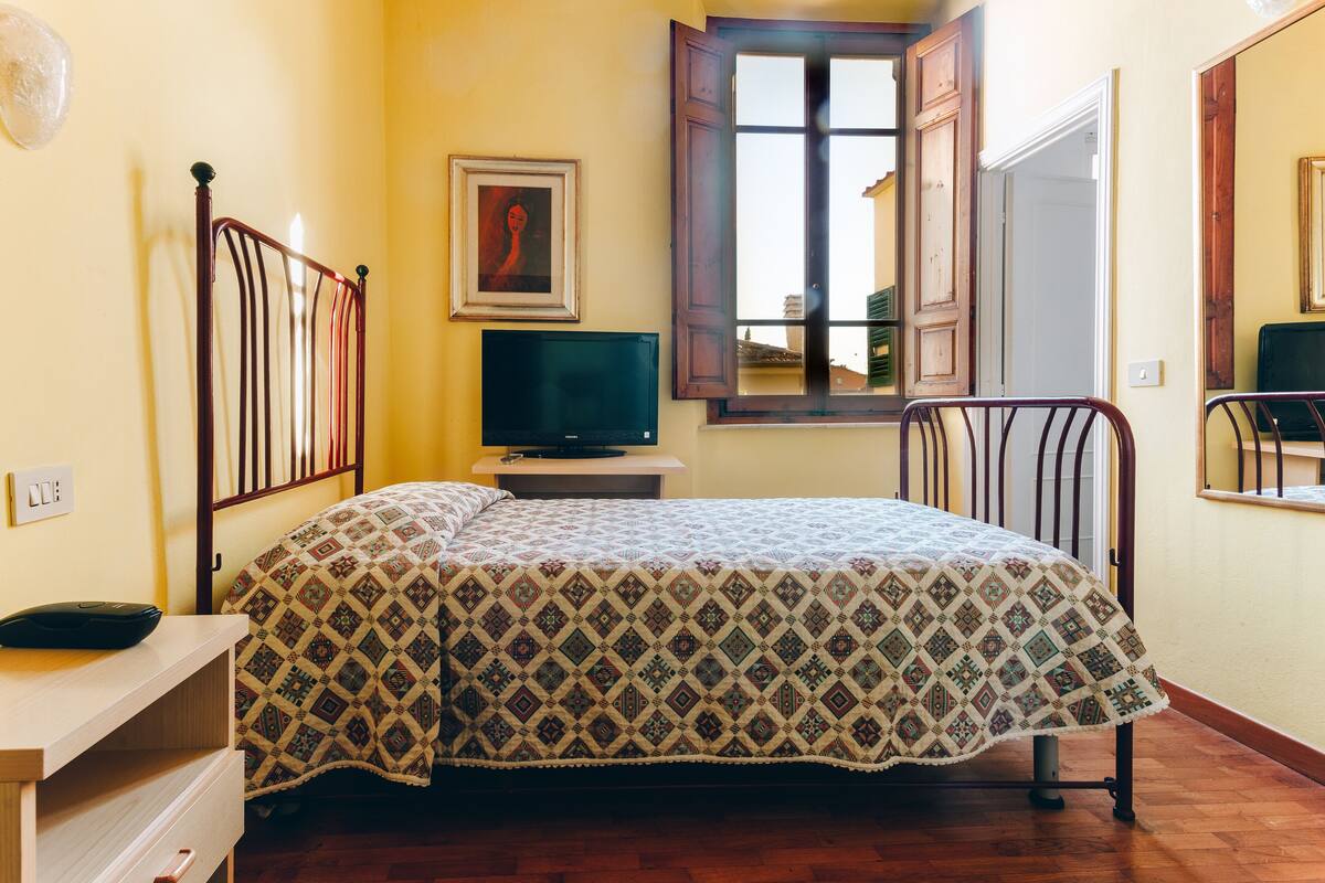 Una habitación con una cama, un televisor sobre un mueble, una ventana con contraventanas de madera y un cuadro en la pared.