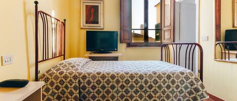 A room with a bed, a television on a stand, a window with wooden shutters, and a painting on the wall.