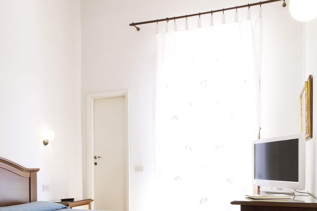 Un dormitorio con cama, escritorio con computadora, silla, puerta y ventana con cortinas.