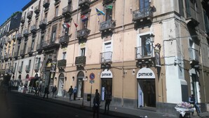 A building with multiple balconies, storefronts, and a street in front. - Rubens (Catania)