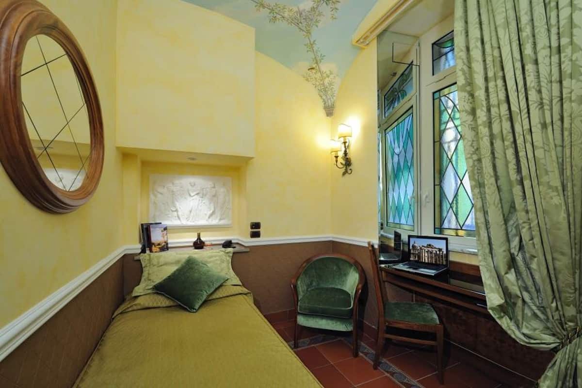Ein Zimmer mit einem Bett, einem Schreibtisch mit Laptop, einem Stuhl, einem Fenster mit Vorhängen und einer Wandlampe.
