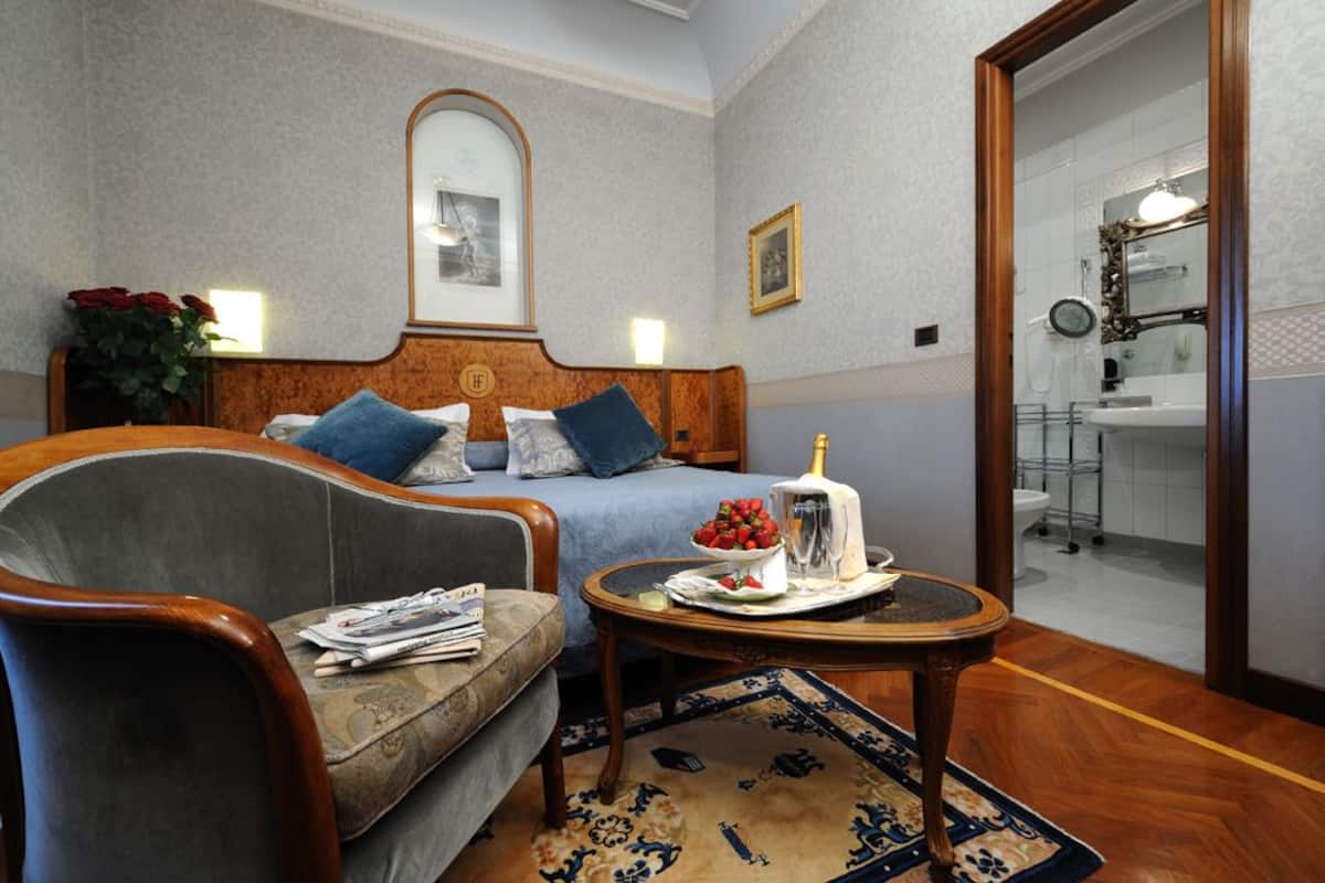 Ein Hotelzimmer mit Bett, einer Couch, einem kleinen Tisch mit Obstkorb und einer Flasche, einem Spiegel und einem durch eine offene Tür einsehbaren Badezimmer.