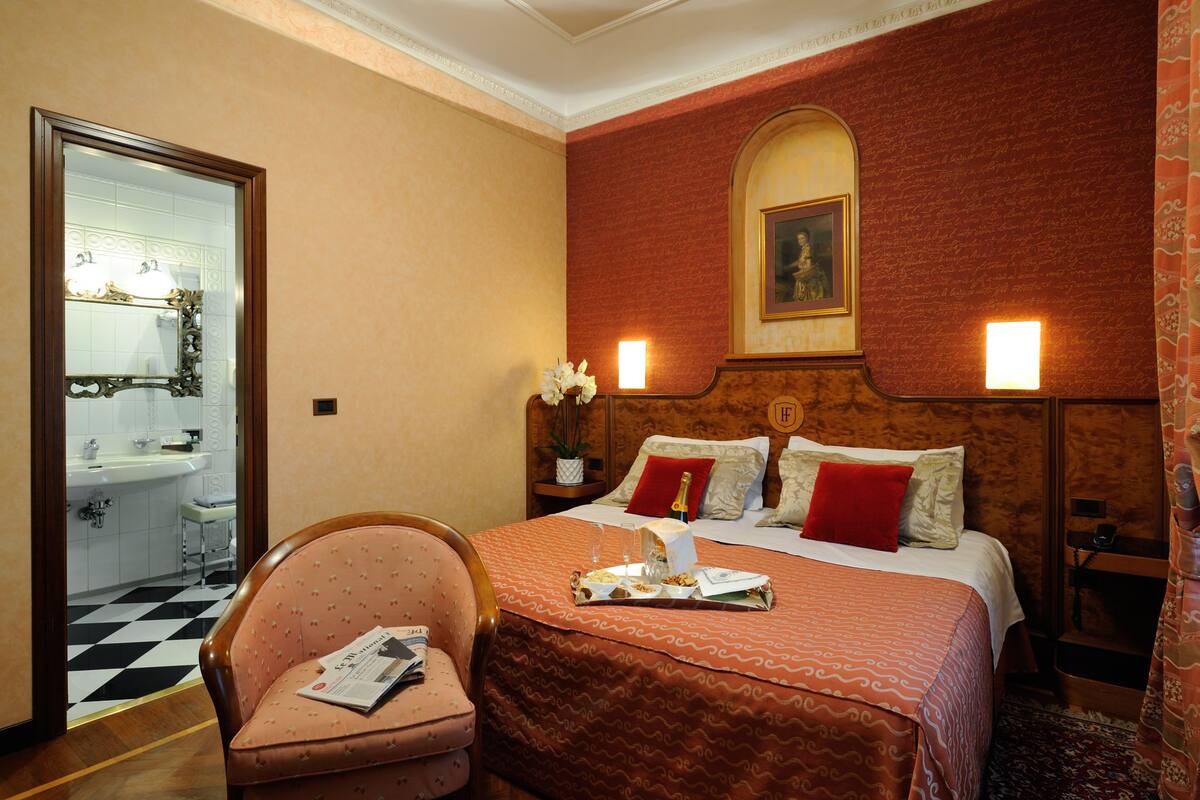 Ein Hotelzimmer mit einem Bett, einem Sessel, einem Nachttisch, einem Spiegel, einem Waschbecken und einer Tür zu einem weiteren Raum.