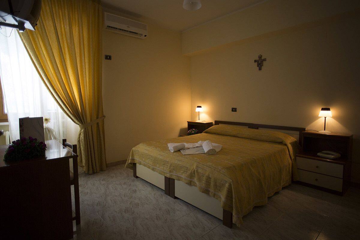 A hotel room with a bed, bedside lamps, a nightstand, a desk with a plant, and a window with curtains.