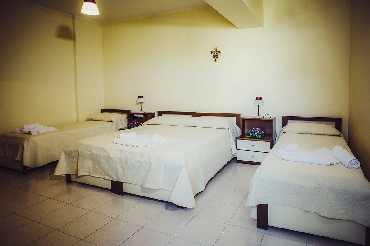 A hotel room with three single beds, each with a white bedspread and a rolled towel. There are bedside tables with lamps and a wall-mounted cross.