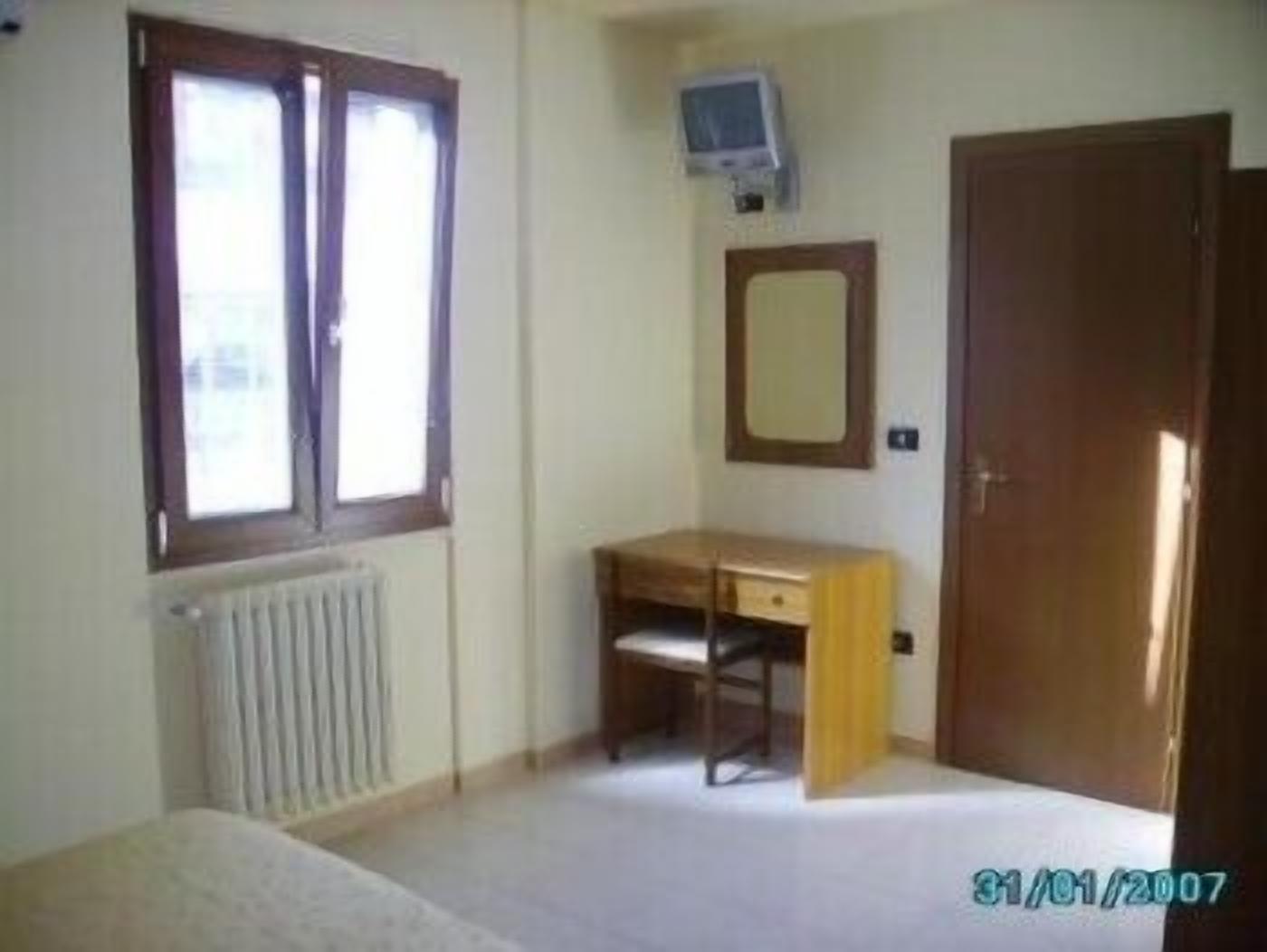 Una stanza piccola e spoglia, con termosifone, una scrivania in legno, una sedia, uno specchio e una porta.