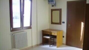 Una stanza piccola e spoglia, con termosifone, una scrivania in legno, una sedia, uno specchio e una porta.