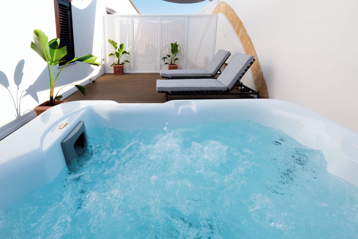 A hot tub with two lounge chairs, potted plants, and a white building with a sloped roof.