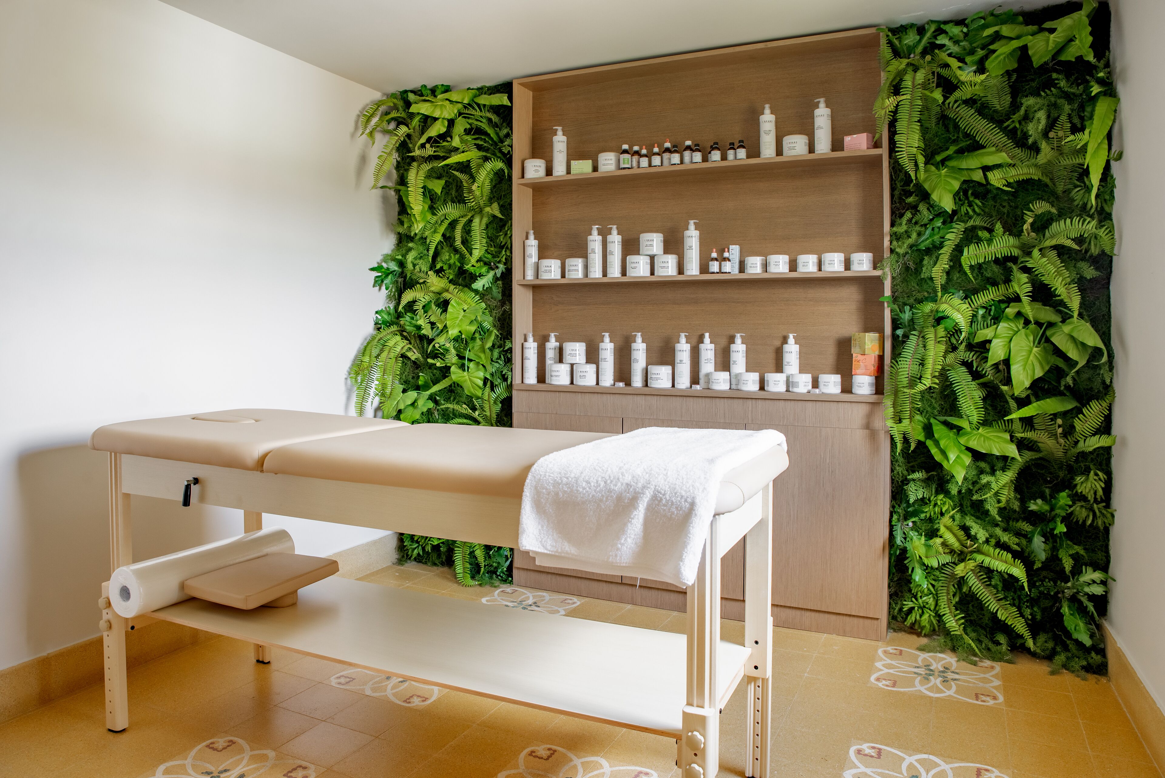 Um espaço de tratamentos com uma estante de produtos para a pele, um banco com toalha e folhagem verde nas paredes.