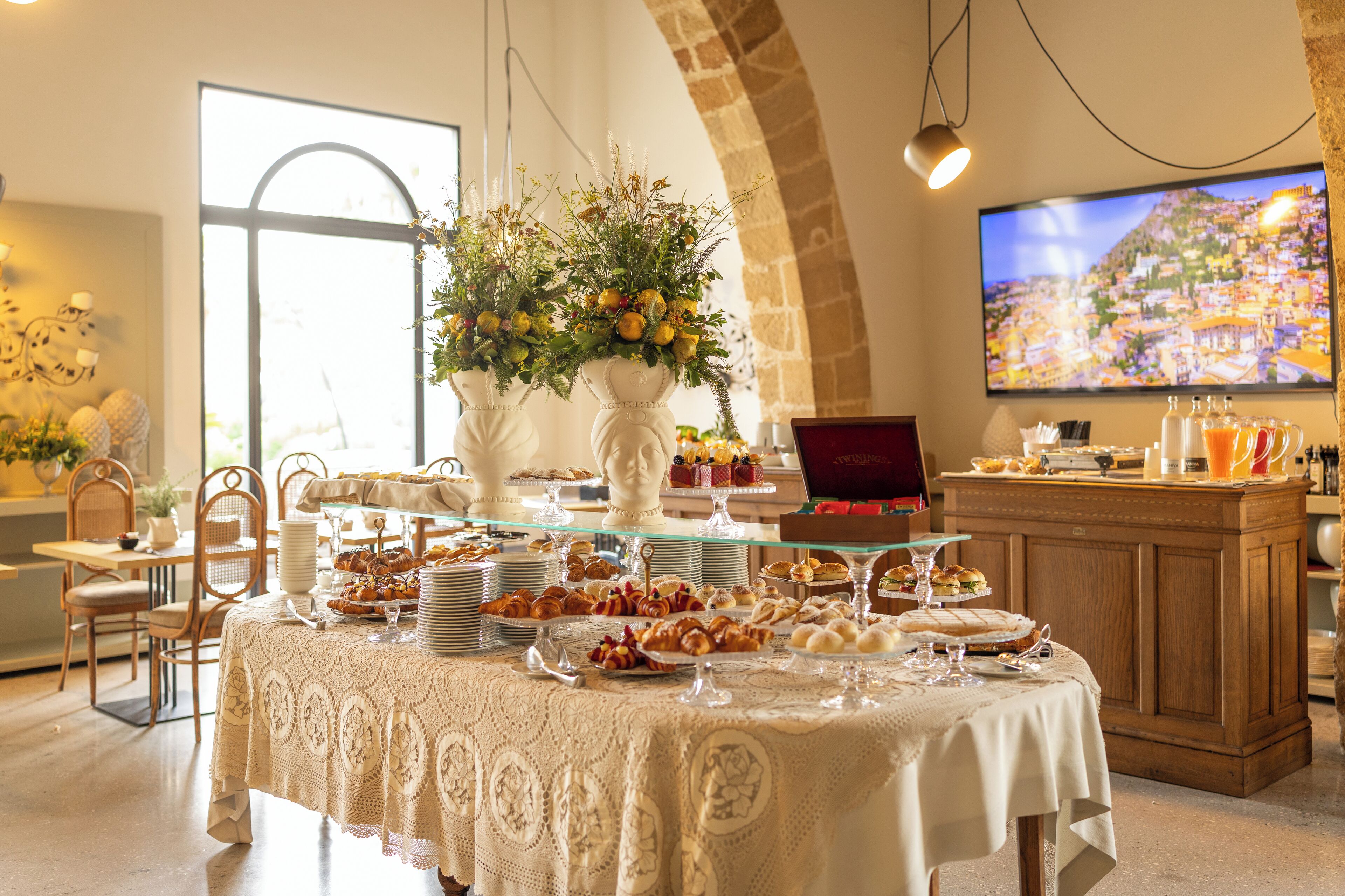 A buffet setup with various food items, a laptop, and a large TV screen in a well-lit room with a stone arch and a large window.